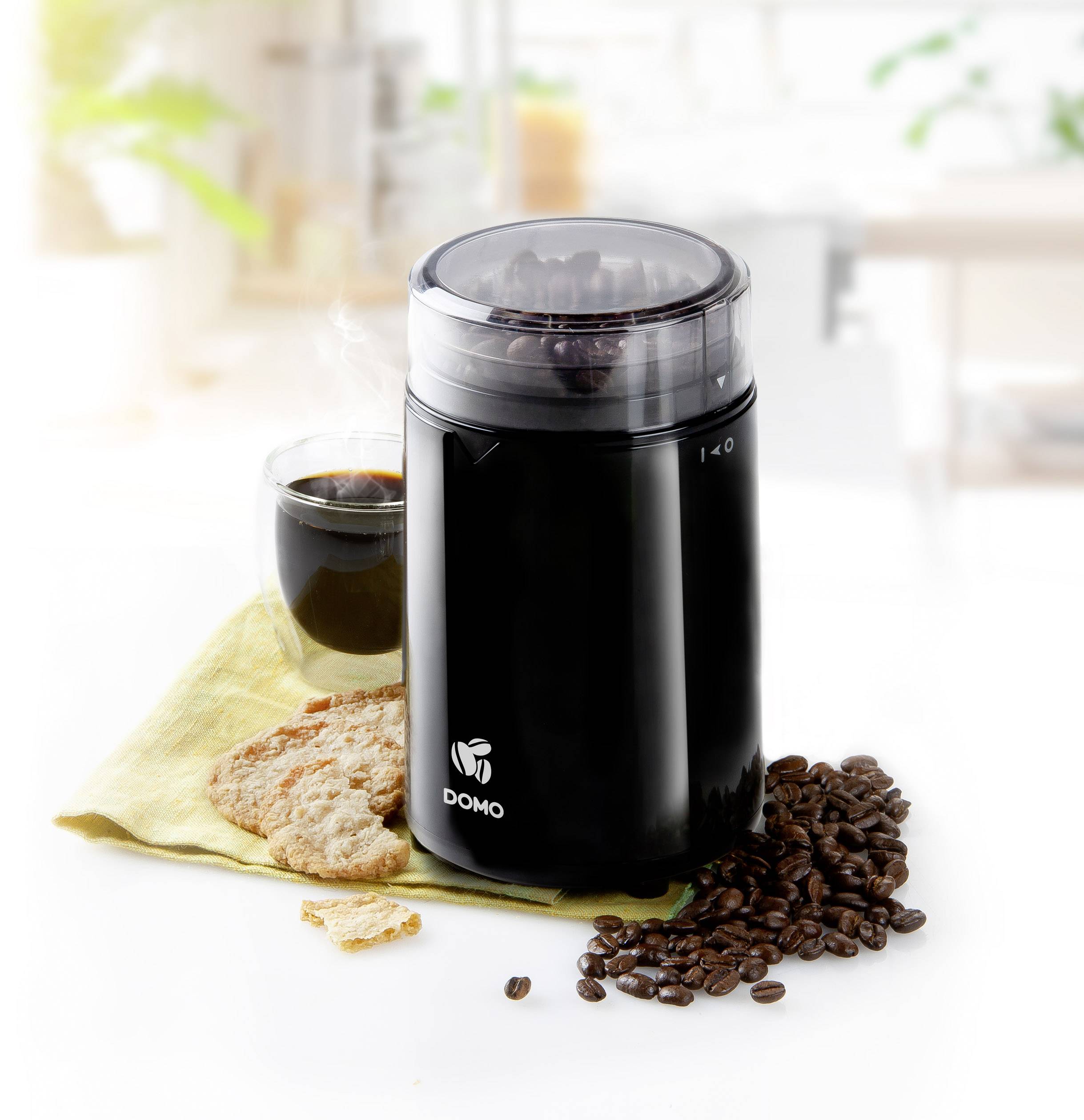 Scene shows a black coffee grinder next to a coffee cup, biscuits and scattered coffee beans on a cloth in a bright kitchen.