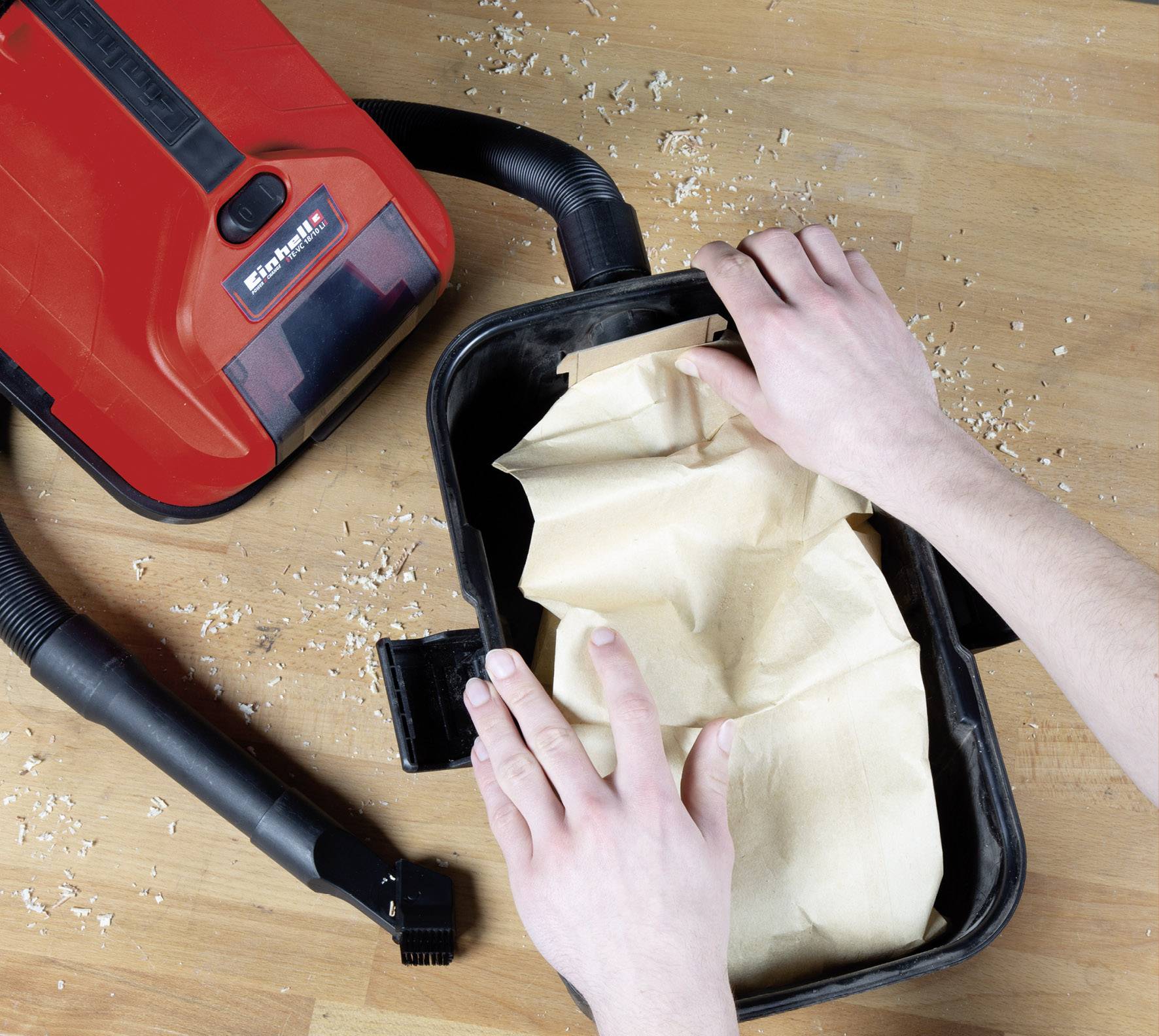 A red industrial vacuum cleaner on a wooden surface; hands are changing a dust bag.