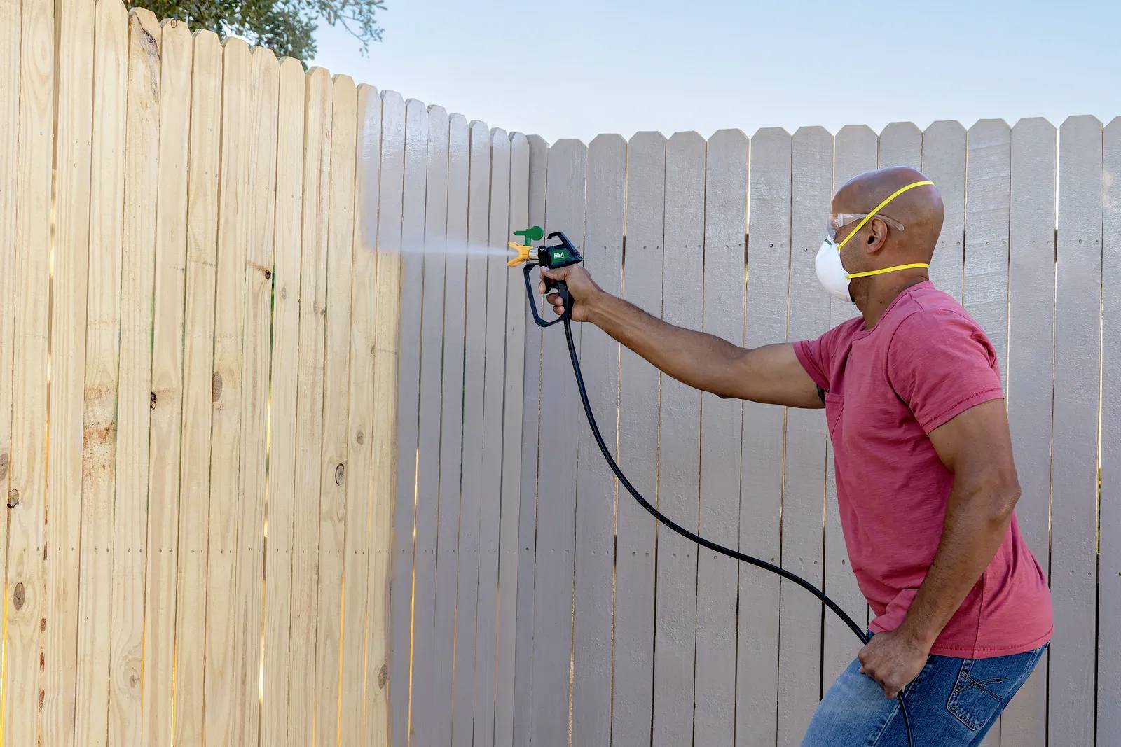 A man wearing a respiratory mask is spraying paint onto a wooden fence.
