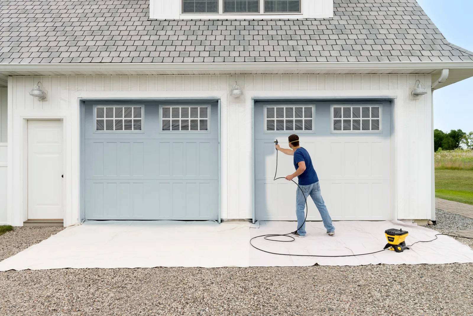 A man is spraying two garage doors with a paint sprayer. The garage doors are light blue, and the house has white walls.