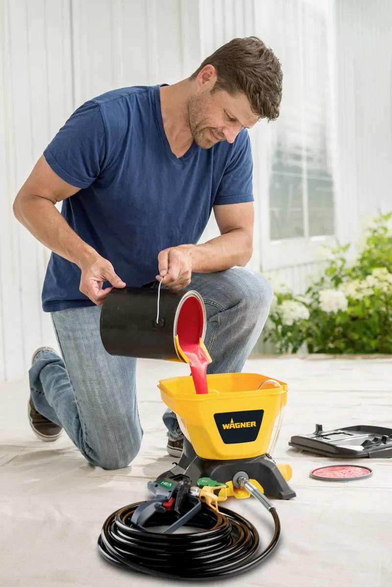 A man is mixing coloured liquid in a yellow Wagner paint spraying system. Tools and hoses are visible on the floor.