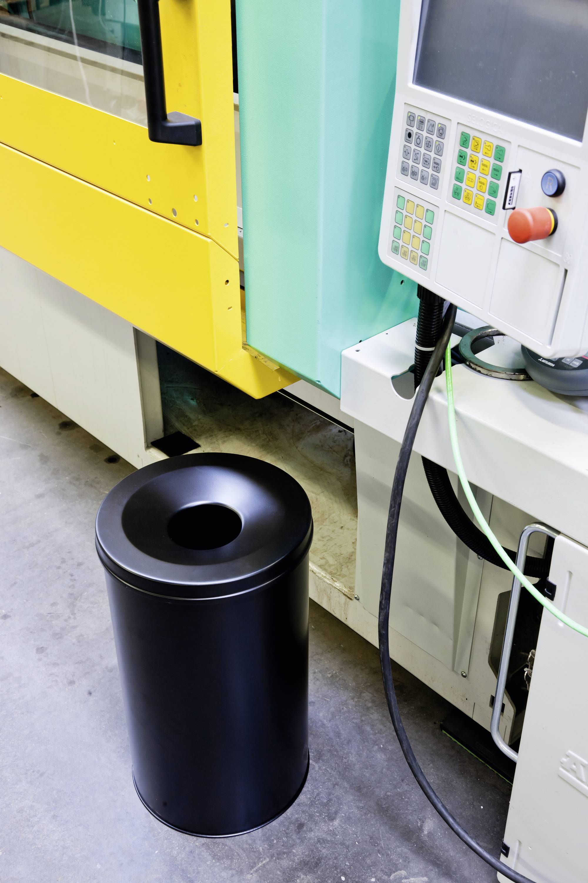 Bin next to a green industrial machine with a yellow protective panel and control panel with buttons and display in a production area.