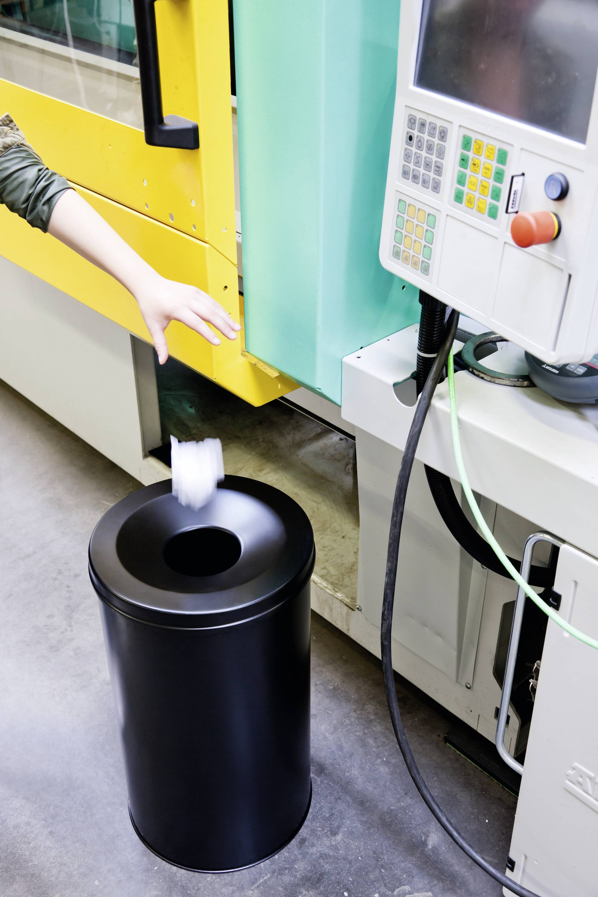 A person throws a crumpled piece of paper into a black rubbish bin next to an industrial machine console in a factory setting.