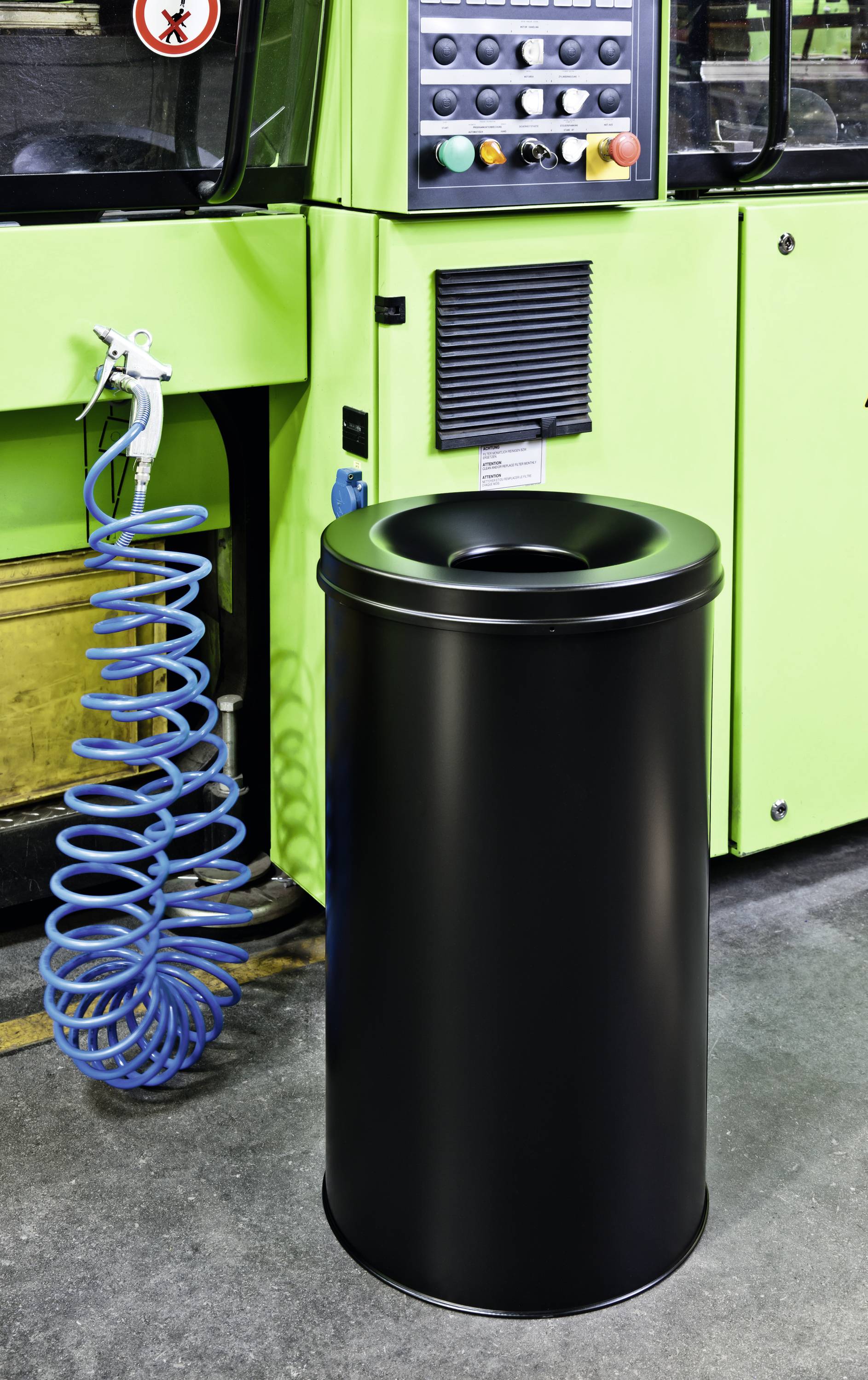 A black dustbin stands in front of a green industrial machine. Beside it, a coiled blue air hose lies on the floor.