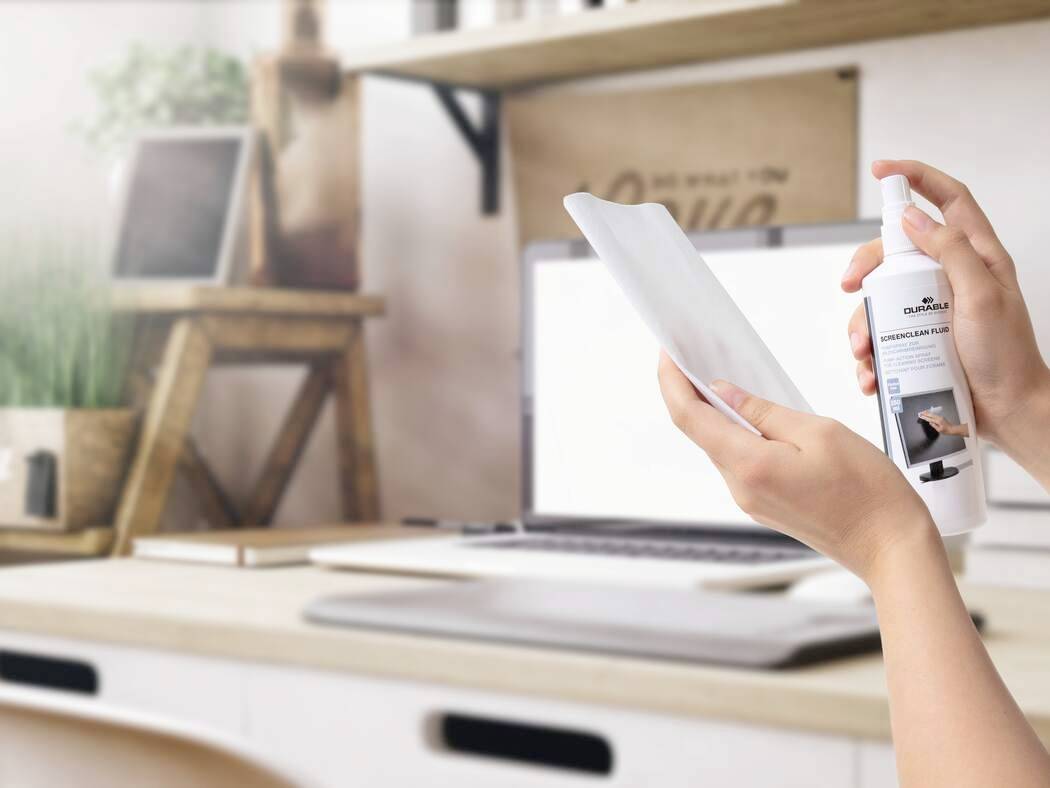 Person cleaning a tablet screen with a spray bottle in a home office setting, featuring a laptop, plant, and shelves in the background.