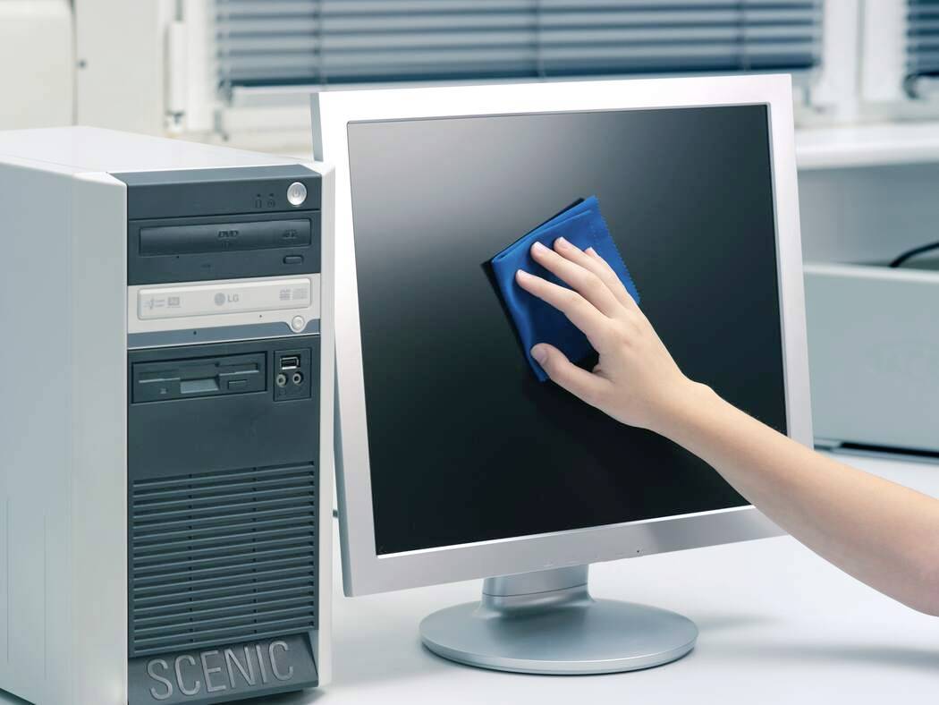 A person cleans a flat-screen computer monitor with a blue cloth in an office setting, next to a desktop CPU.