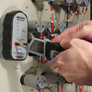A person is using a digital measuring device to check the voltage on an electrical control unit. Several cables are connected.