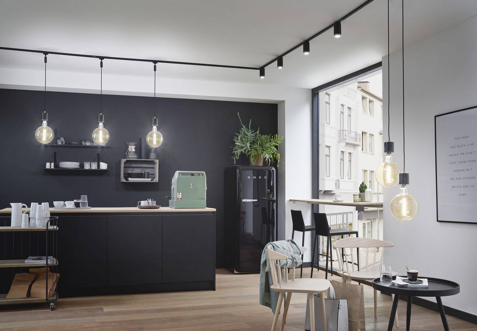 Modern kitchen with black cabinets, wooden worktop and pendant lights. Window overlooking buildings. Plants and coffee machine visible.