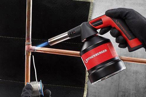 A soldering iron heats a copper pipe. A person holds solder close to the heated area to join the pieces together.