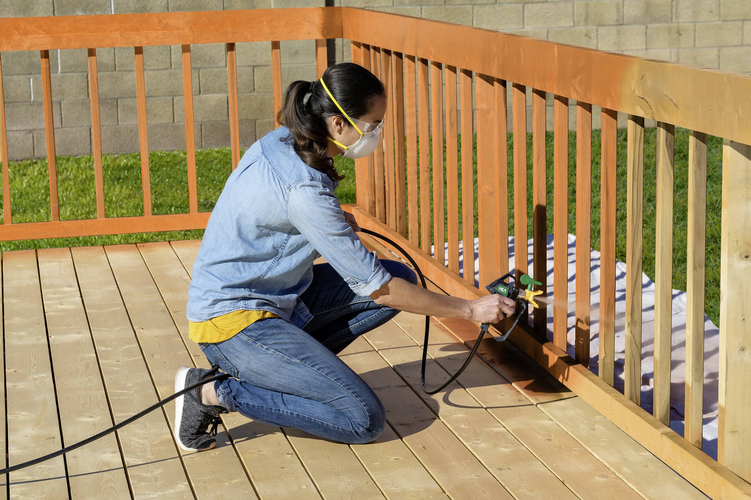 A person is painting a wooden decking with a paint device and is wearing a protective mask for outdoor safety.