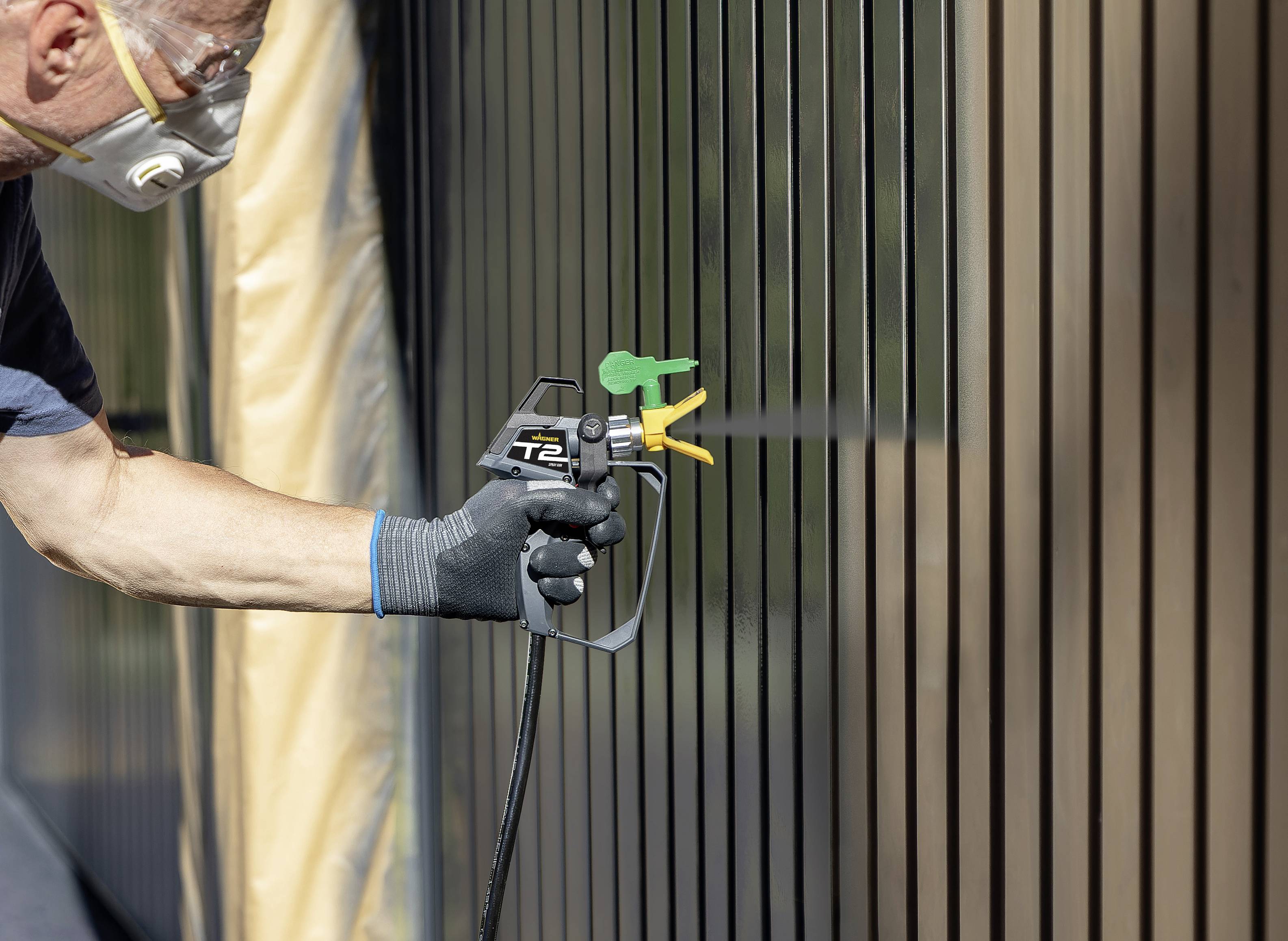 A person is spraying paint onto a fence with a spray gun. They are wearing gloves and a protective mask.