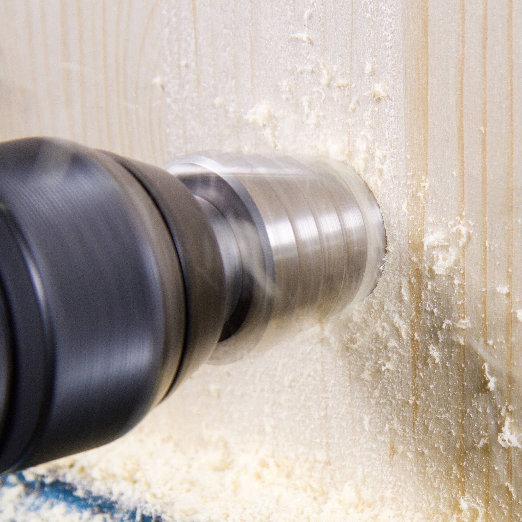 A drill is boring a round hole into a wooden board. Wood shavings are flying around.