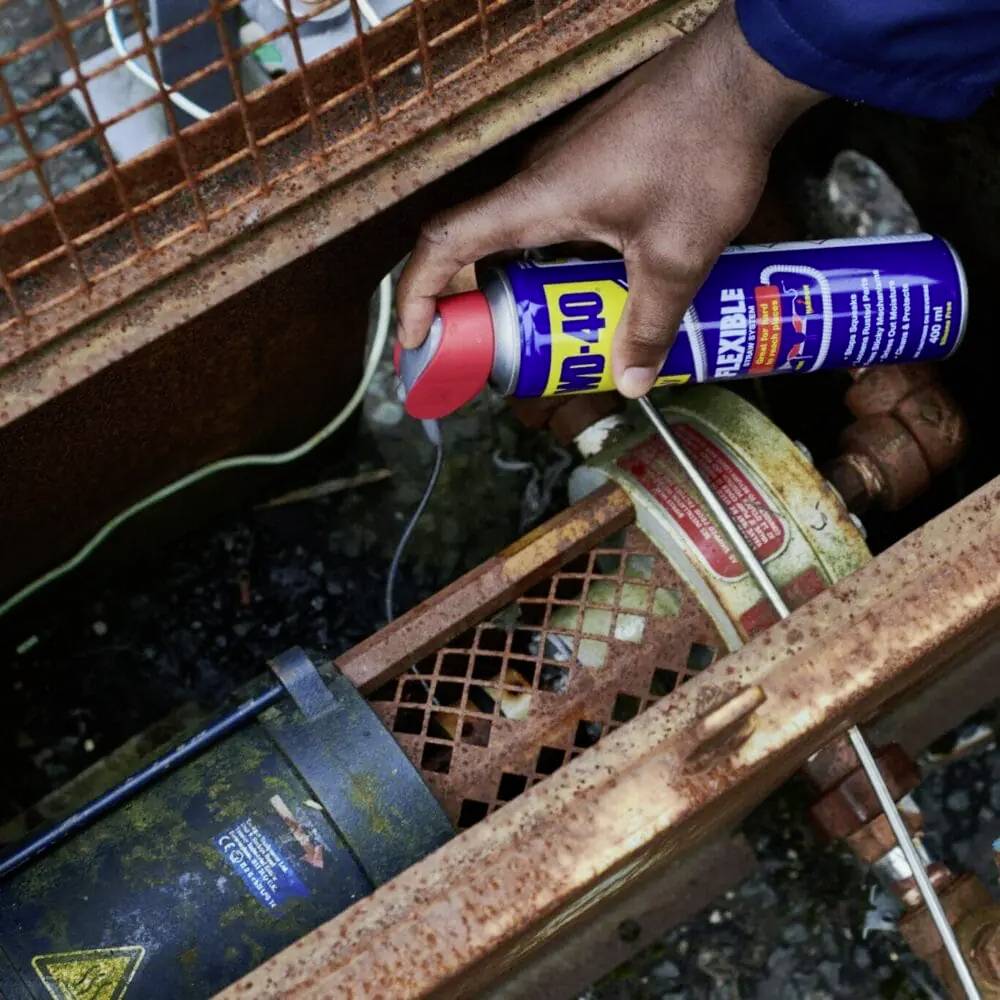A hand holds a blue tin of lubricant and sprays it onto a rusty padlock for maintenance or repair.