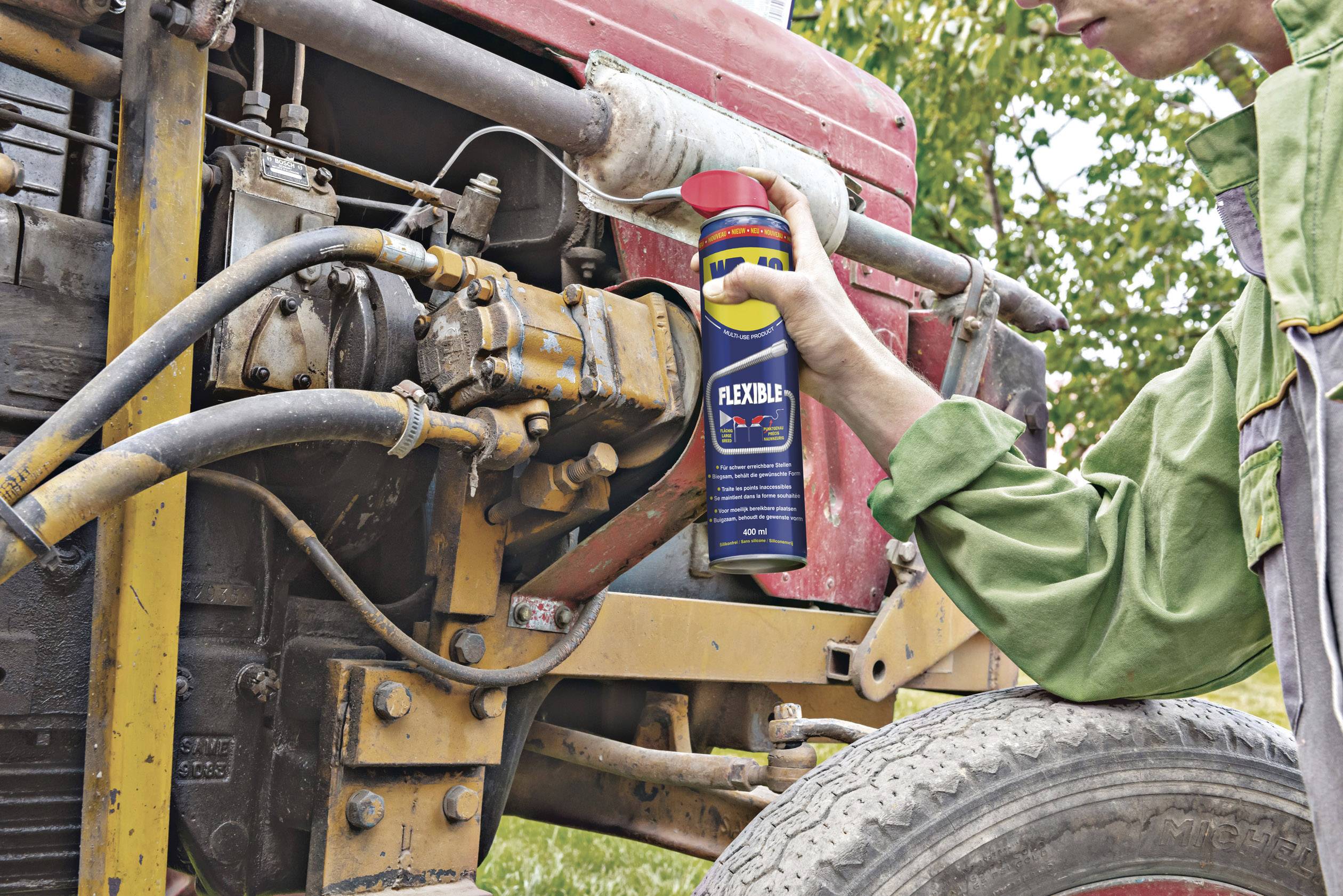 A person is spraying oil onto an old, rusty tractor part. The tractor is standing on a meadow.