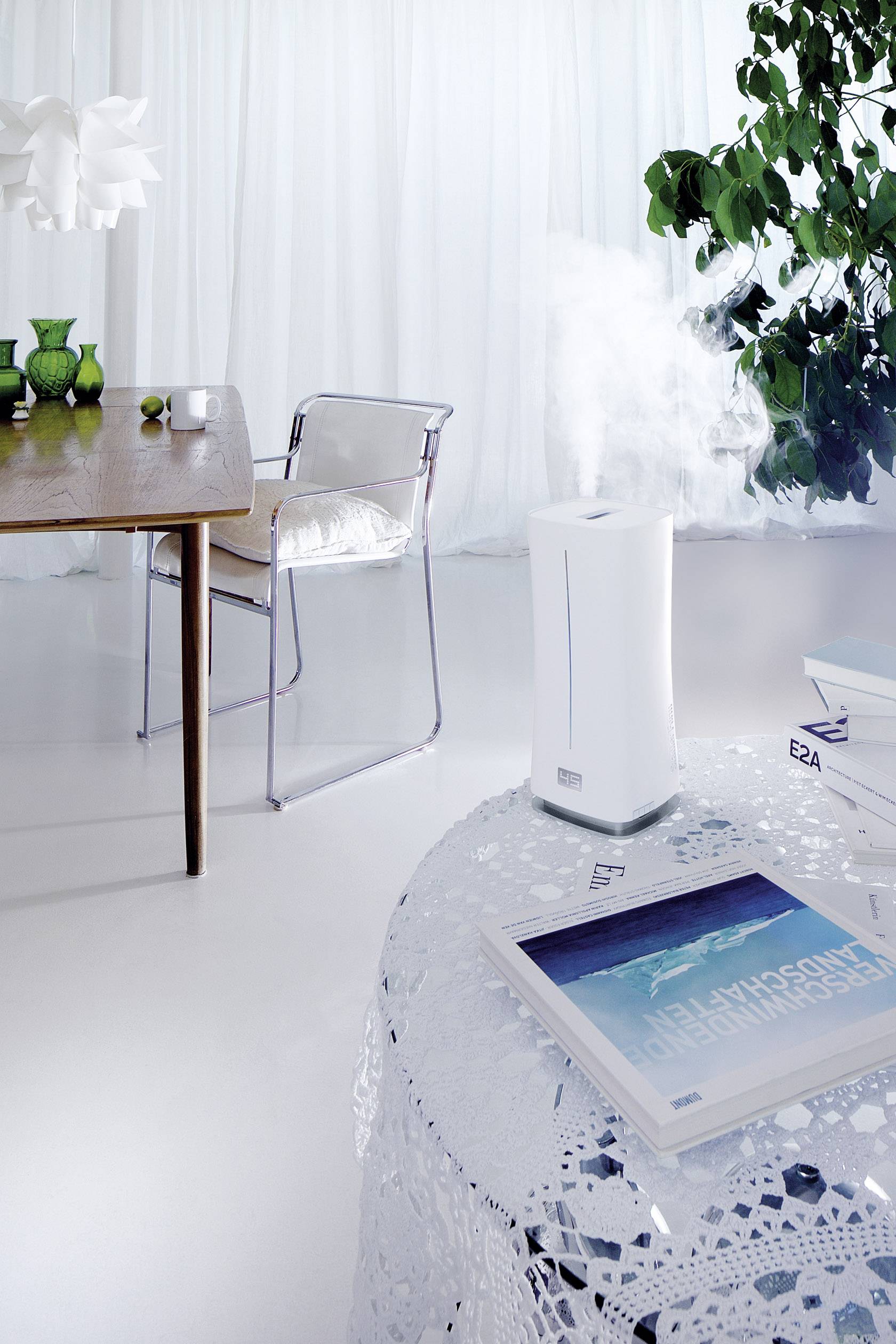 Modern, bright room with a table and chairs, humidifier on table emitting water vapour, plants in the background, light curtains.