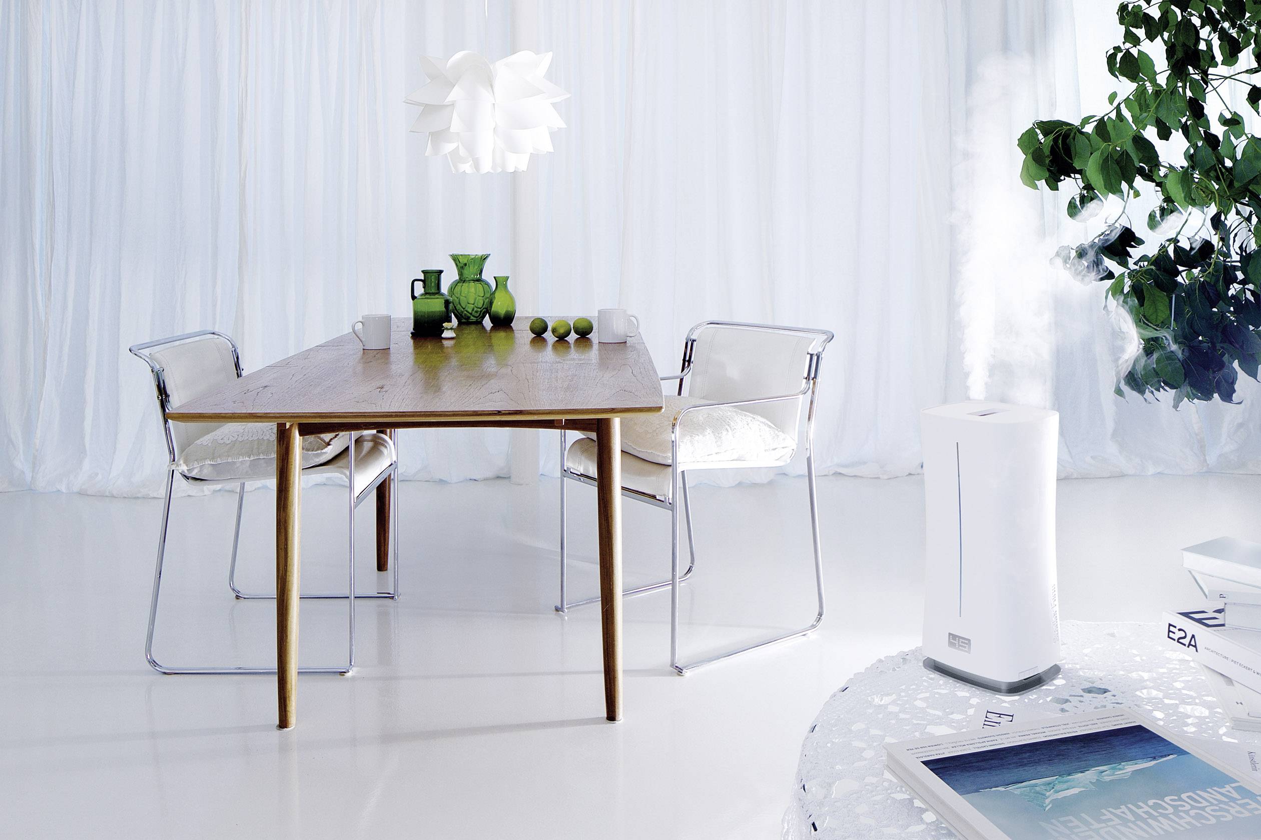 Dining room with a modern wooden table and chairs, decorated with green vases and tumblers. A humidifier is misting water, with botanical decorations in the background.