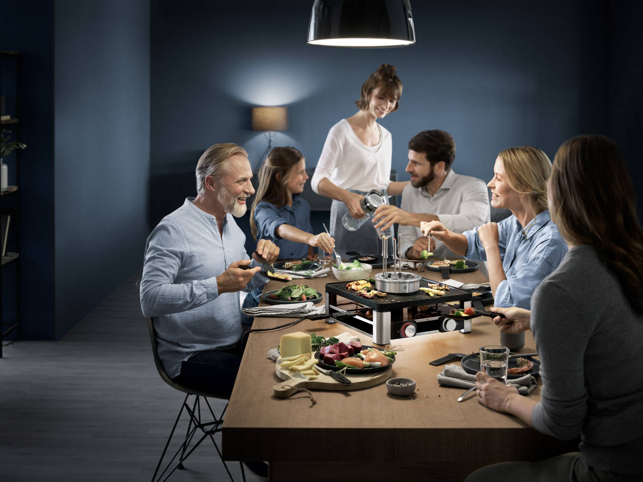 A group of six people are sitting smiling around a table, enjoying a shared meal in a cosy, dimly lit environment.