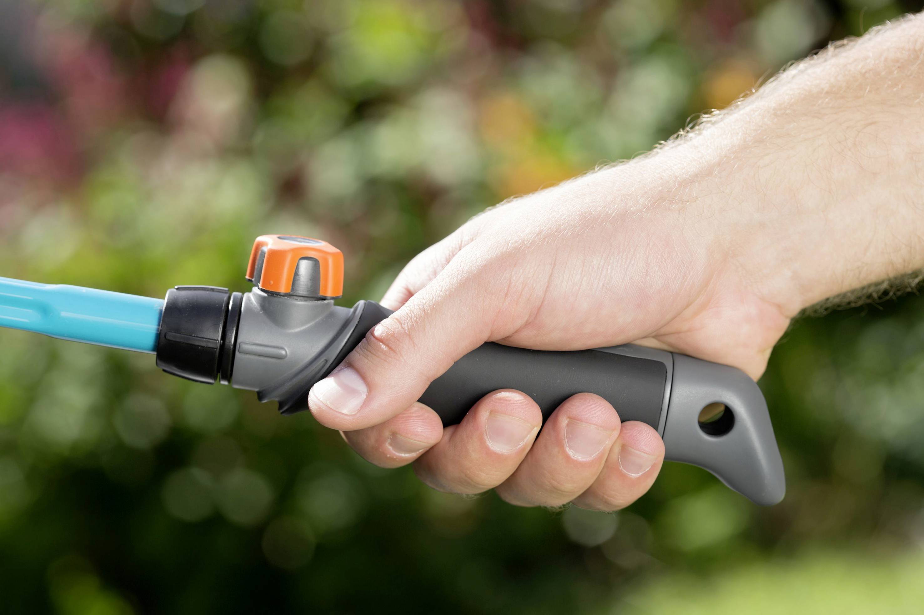 A hand is holding a garden hose with a grey handle and an orange valve against a blurred background.