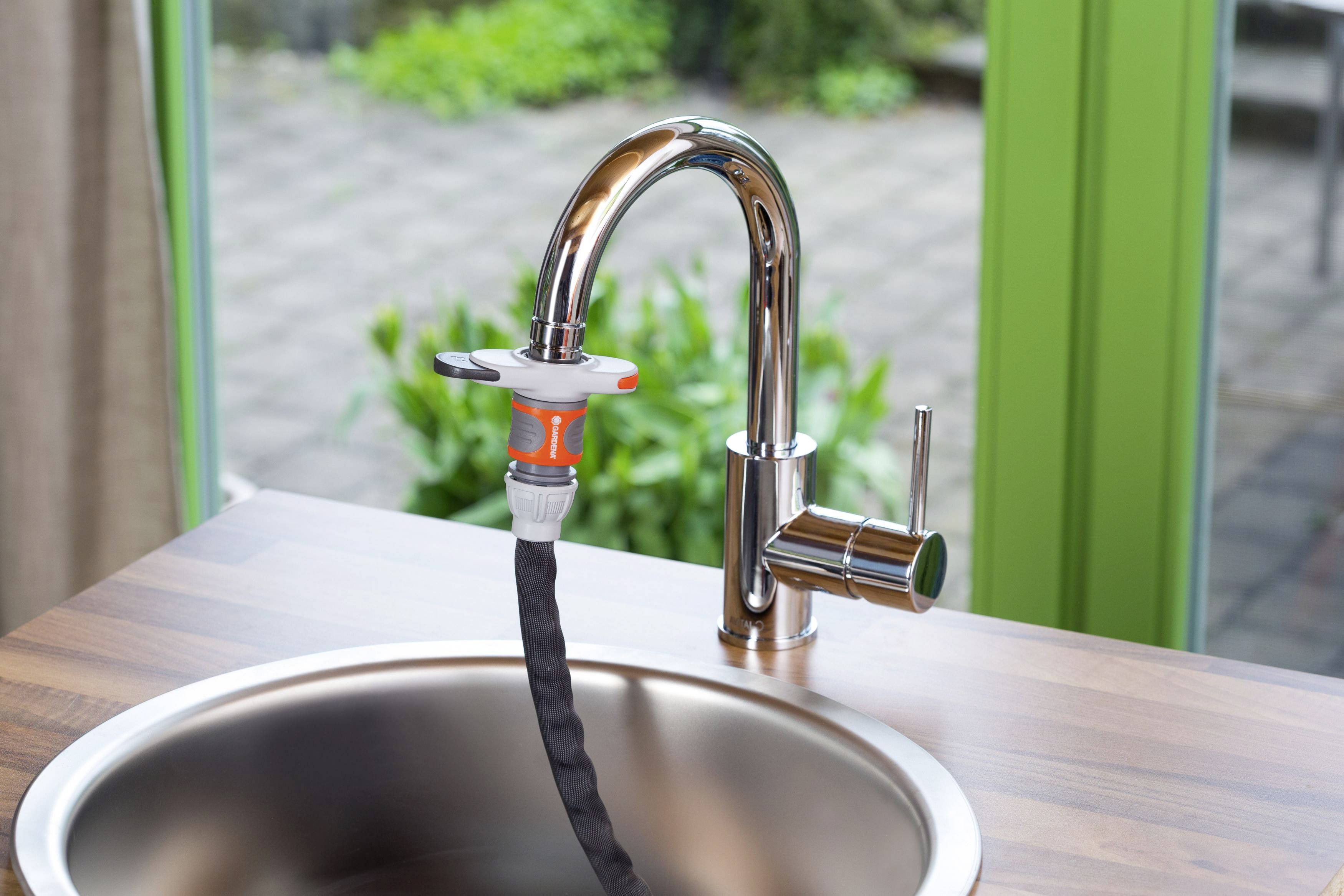 A tap in a kitchen with a garden hose adapter attached. Garden tiles and green plants are visible in the background.