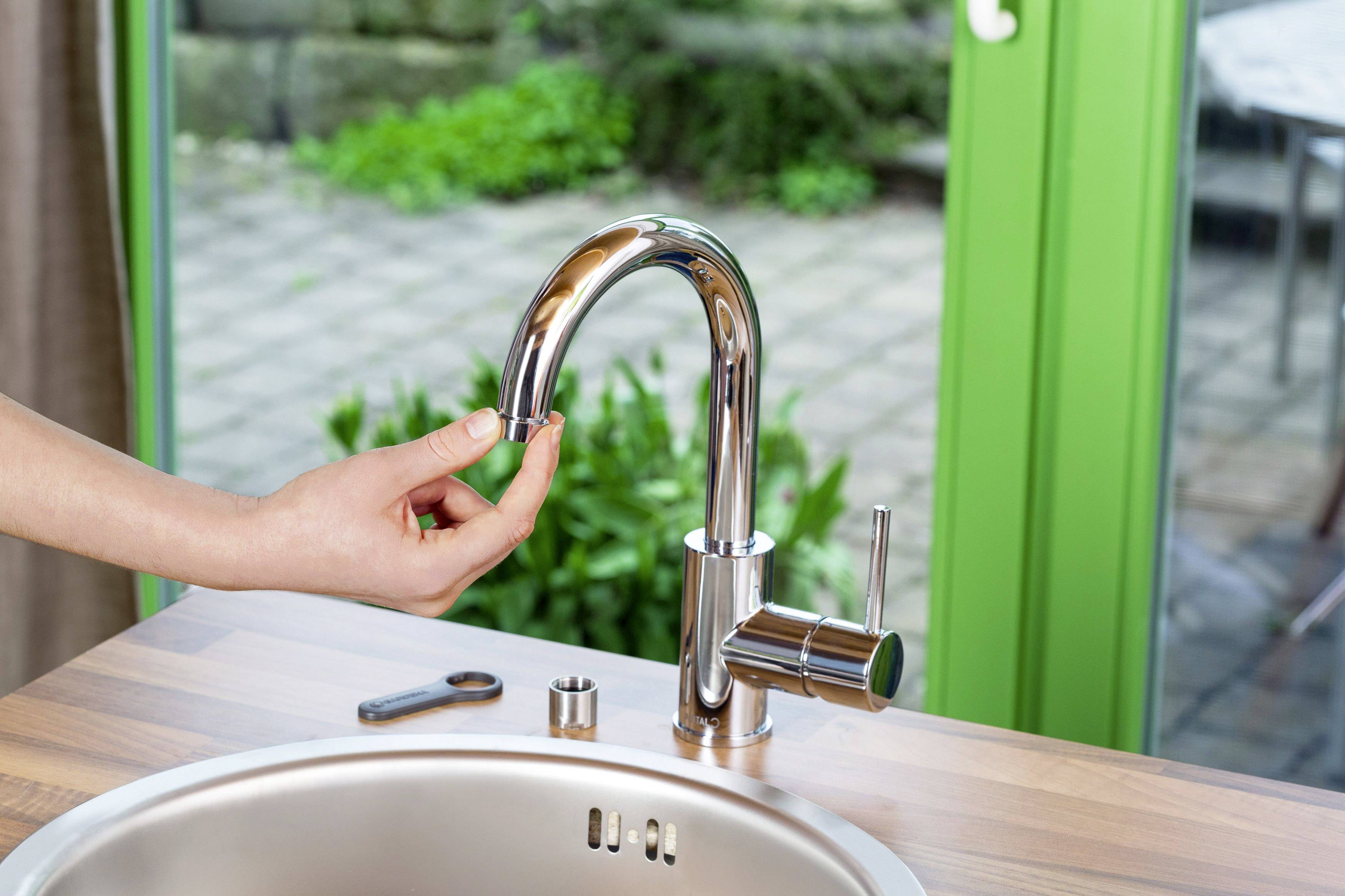 A person is repairing a tap in the kitchen. The tap is made of shiny metal and stands on a wooden worktop.