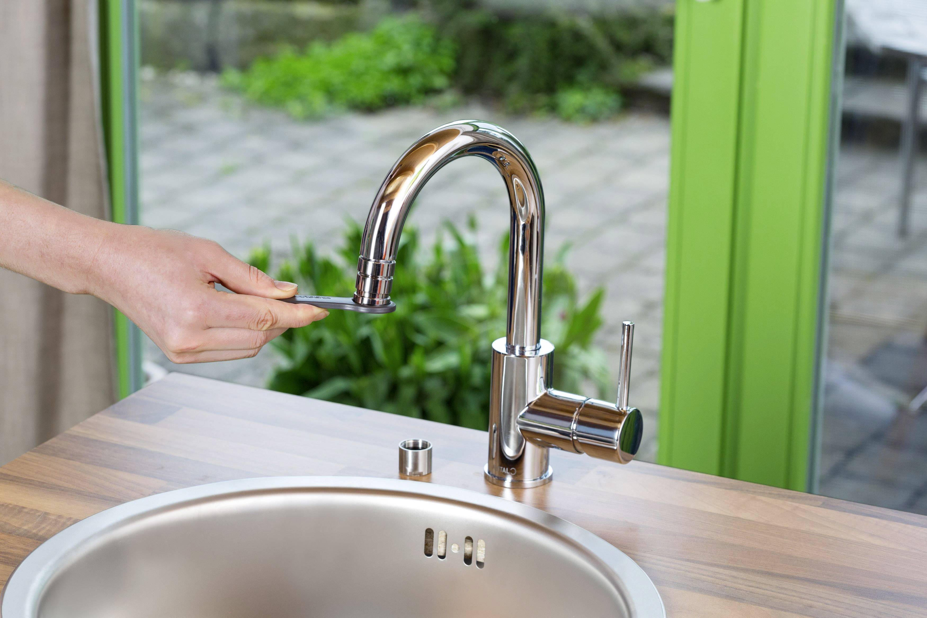 A hand operates the lever of a modern kitchen tap on a wooden worktop. In the background, a window offers a view of a garden.