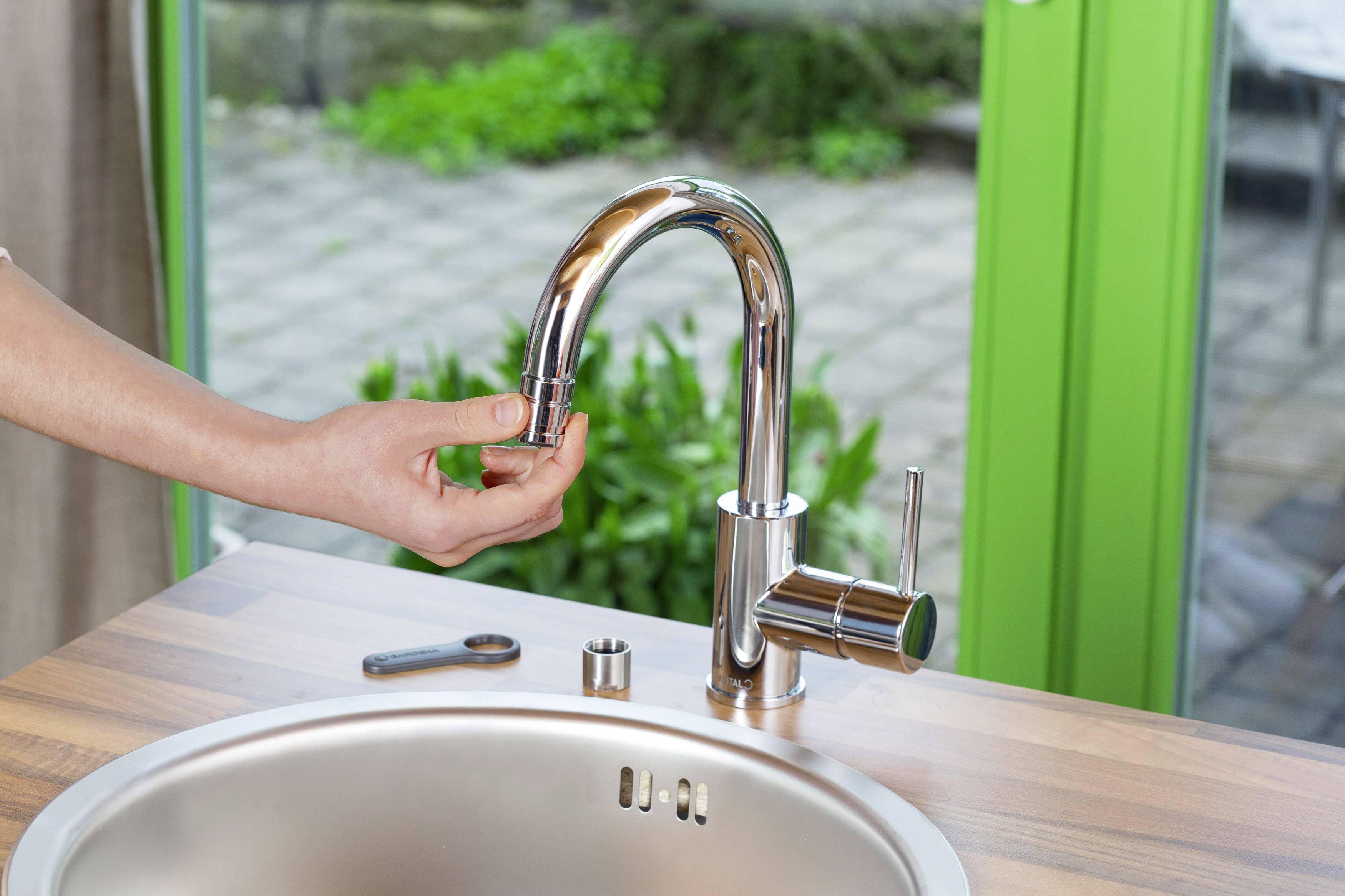 A hand loosens the spray control of a kitchen tap at a sink, with a green garden visible in the background.