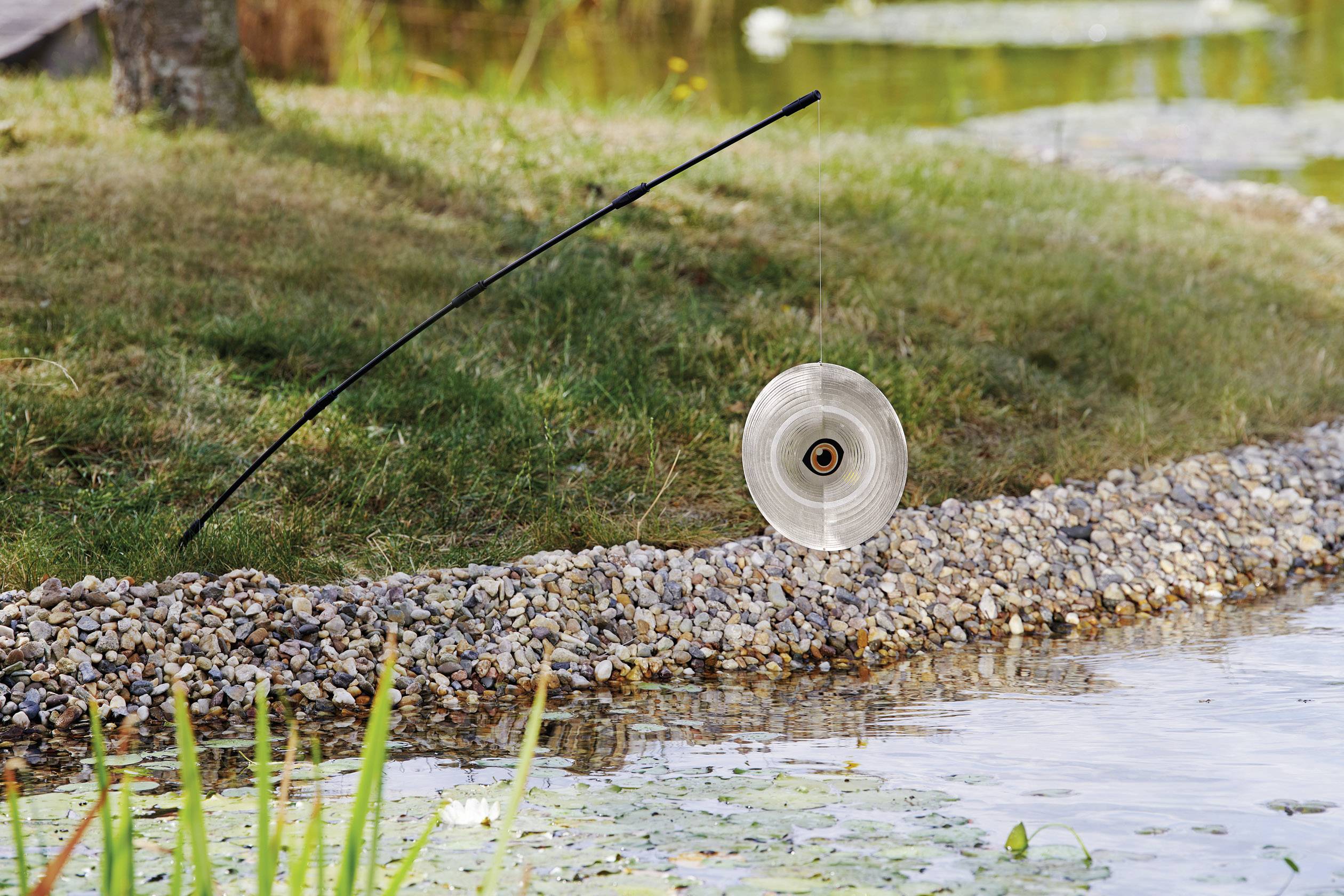 A standing garden hose holder with a coiled hose on a stony bank beside a pond with water lilies.