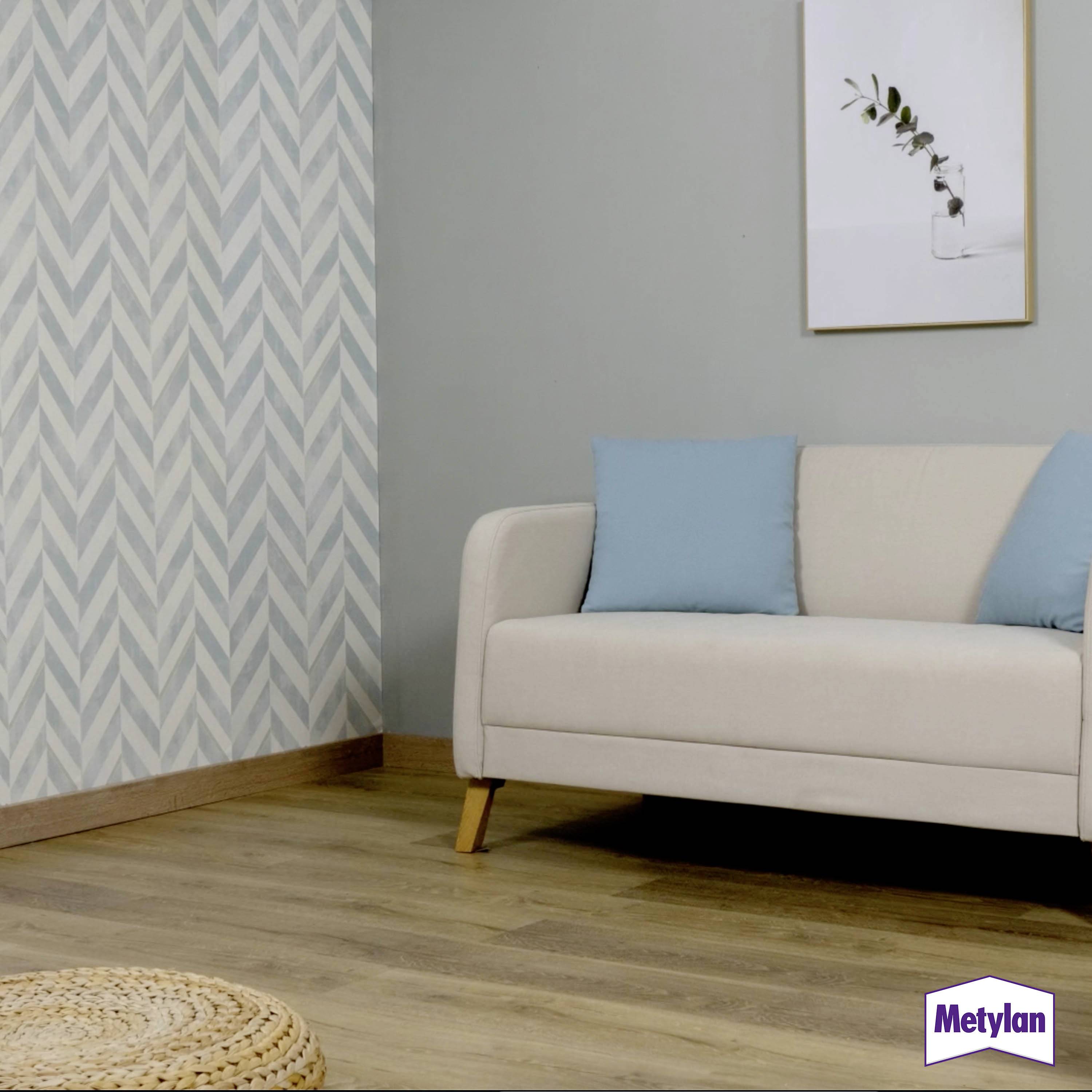 A living room with a beige sofa, blue cushions, light wood floor, and blue-grey patterned wallpaper. A framed plant photo hangs on the wall.