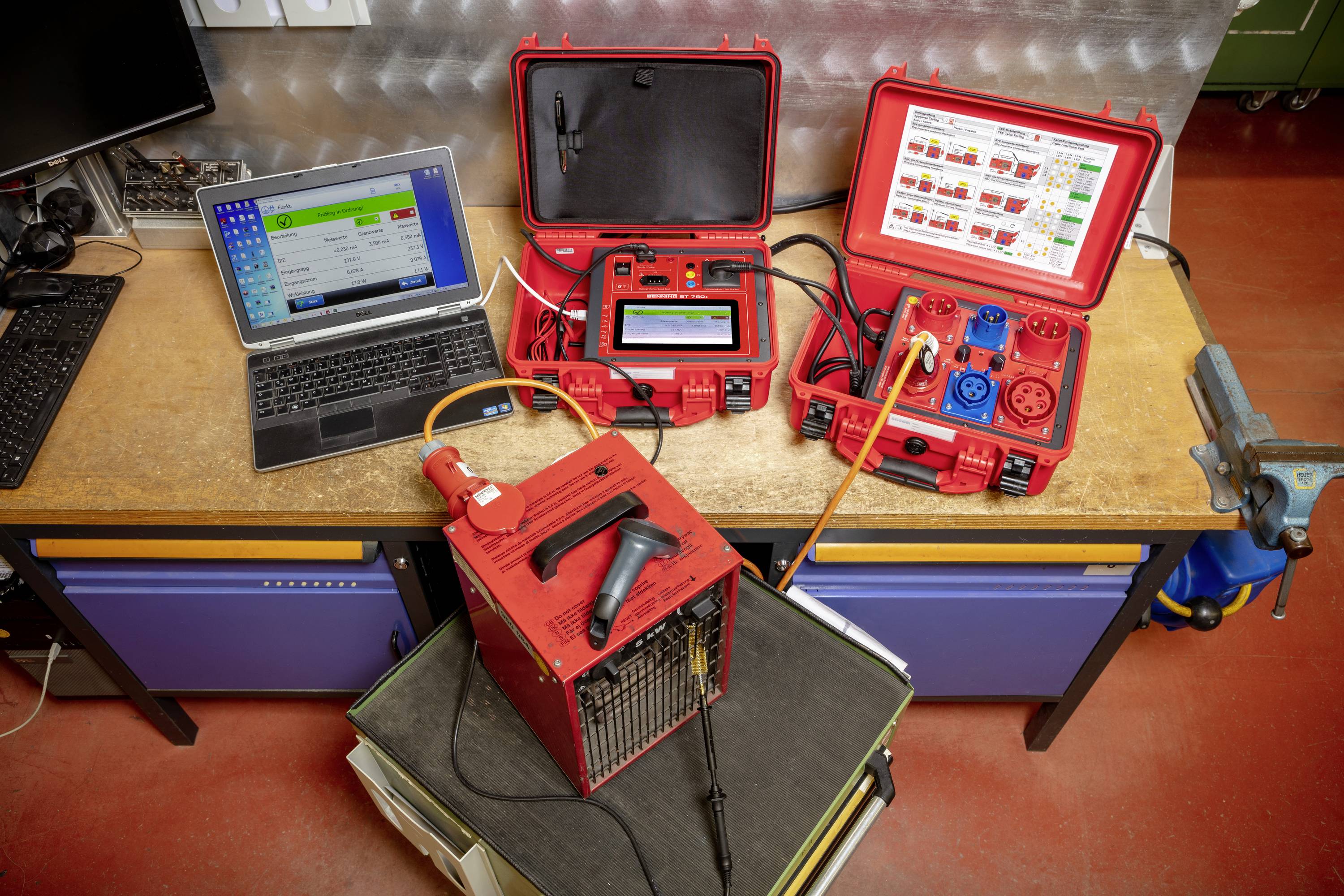 A workshop workstation shows a laptop, two red devices for electrical testing, and a fan heater on a table.