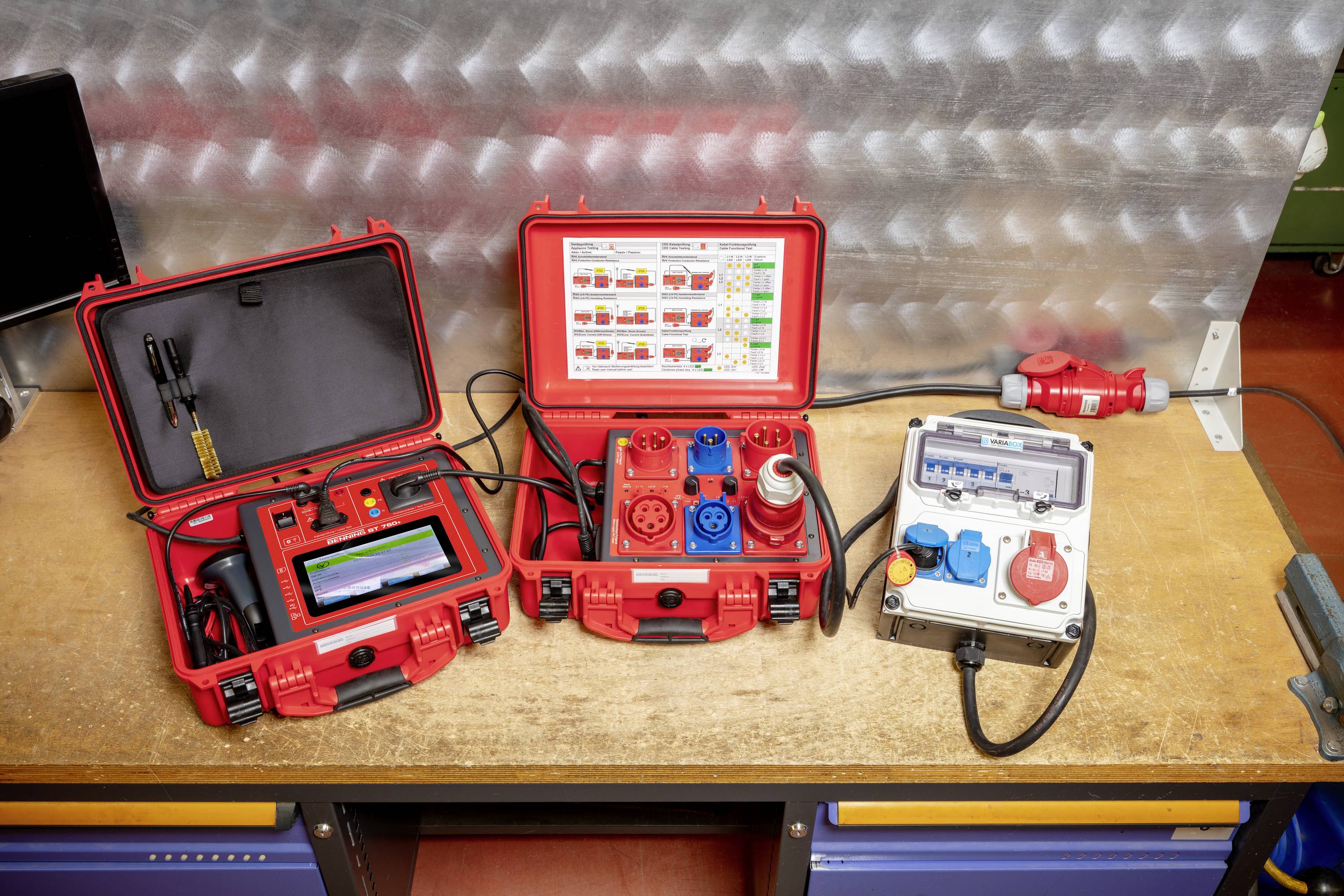 Three red suitcases and a control box with electrical measuring equipment on a table. A screen on the left, plug slots in the centre, power connections on the right.