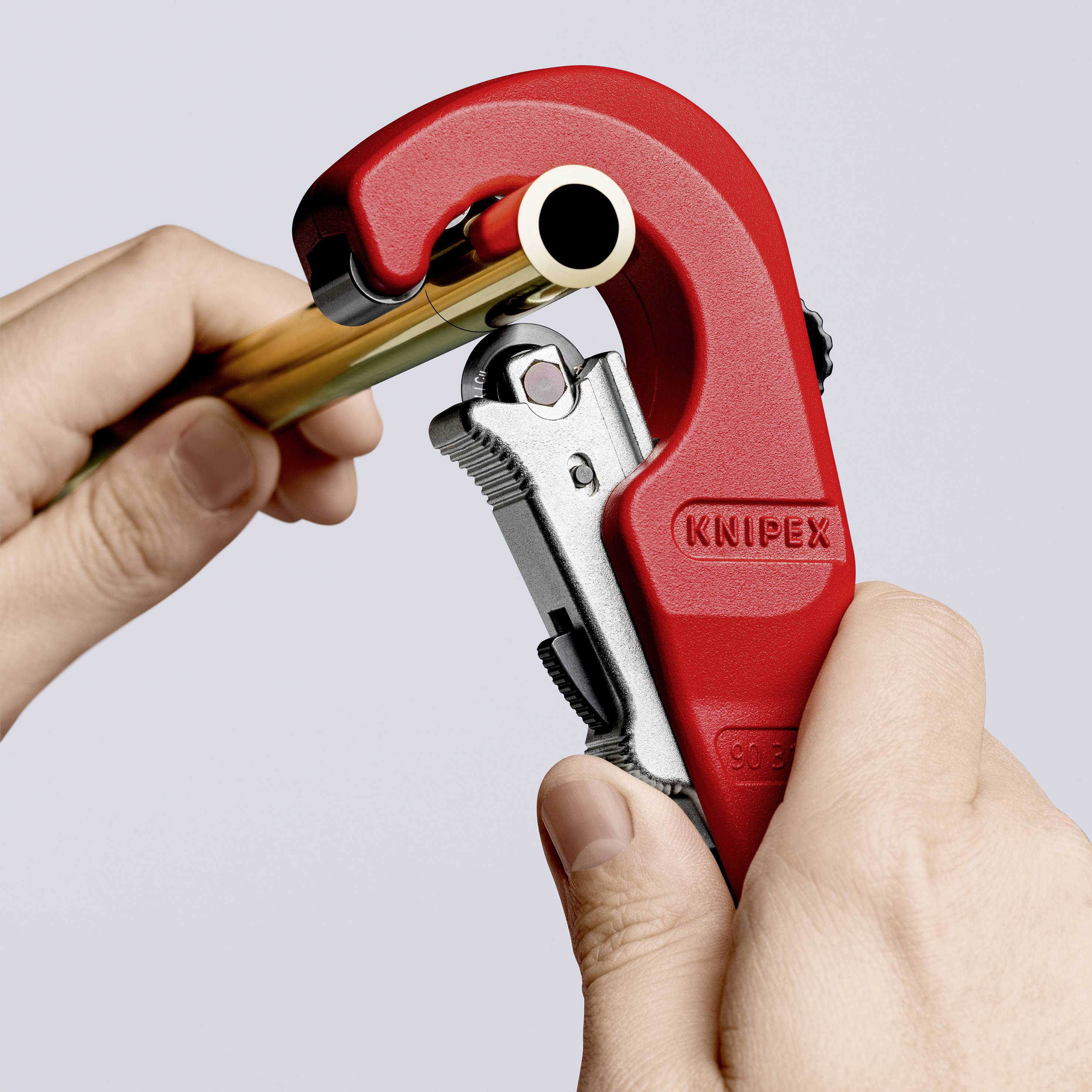 A hand is holding a red pipe cutting tool that is cutting through a metal pipe.