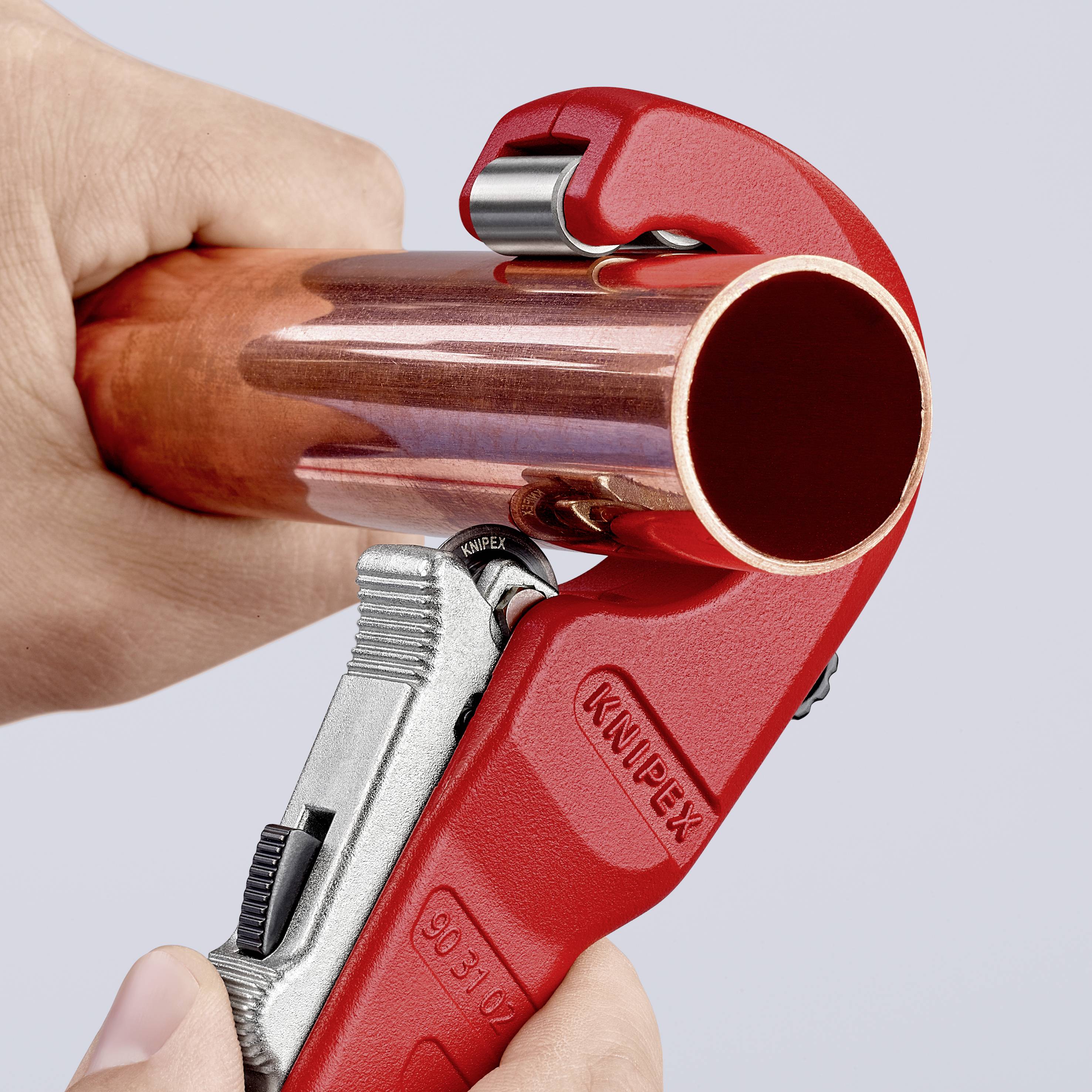 A hand is cutting a copper pipe with a red pipe cutter.