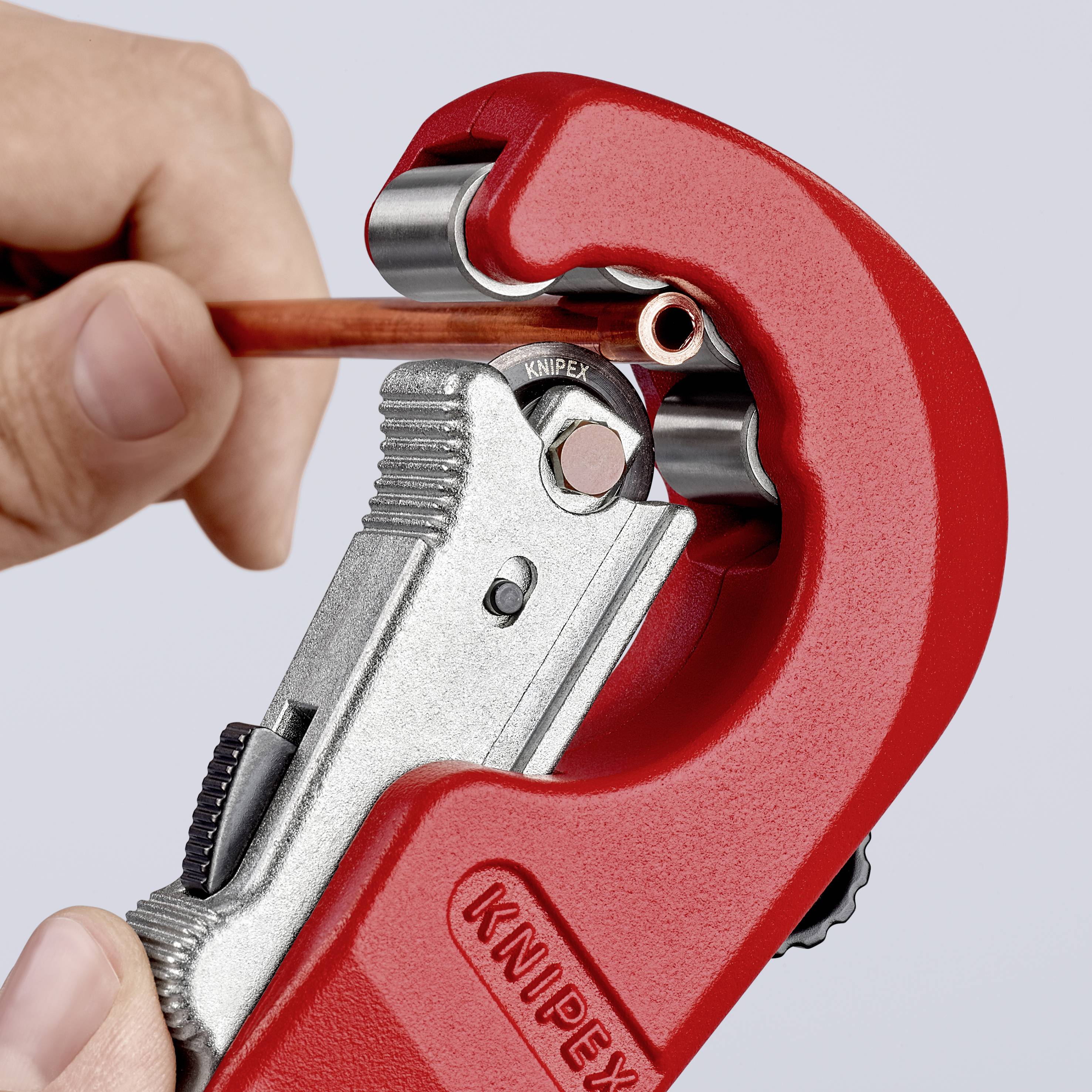 A hand is using a red pipe cutter to cut a copper pipe. The cutter is metallic with a black handle.