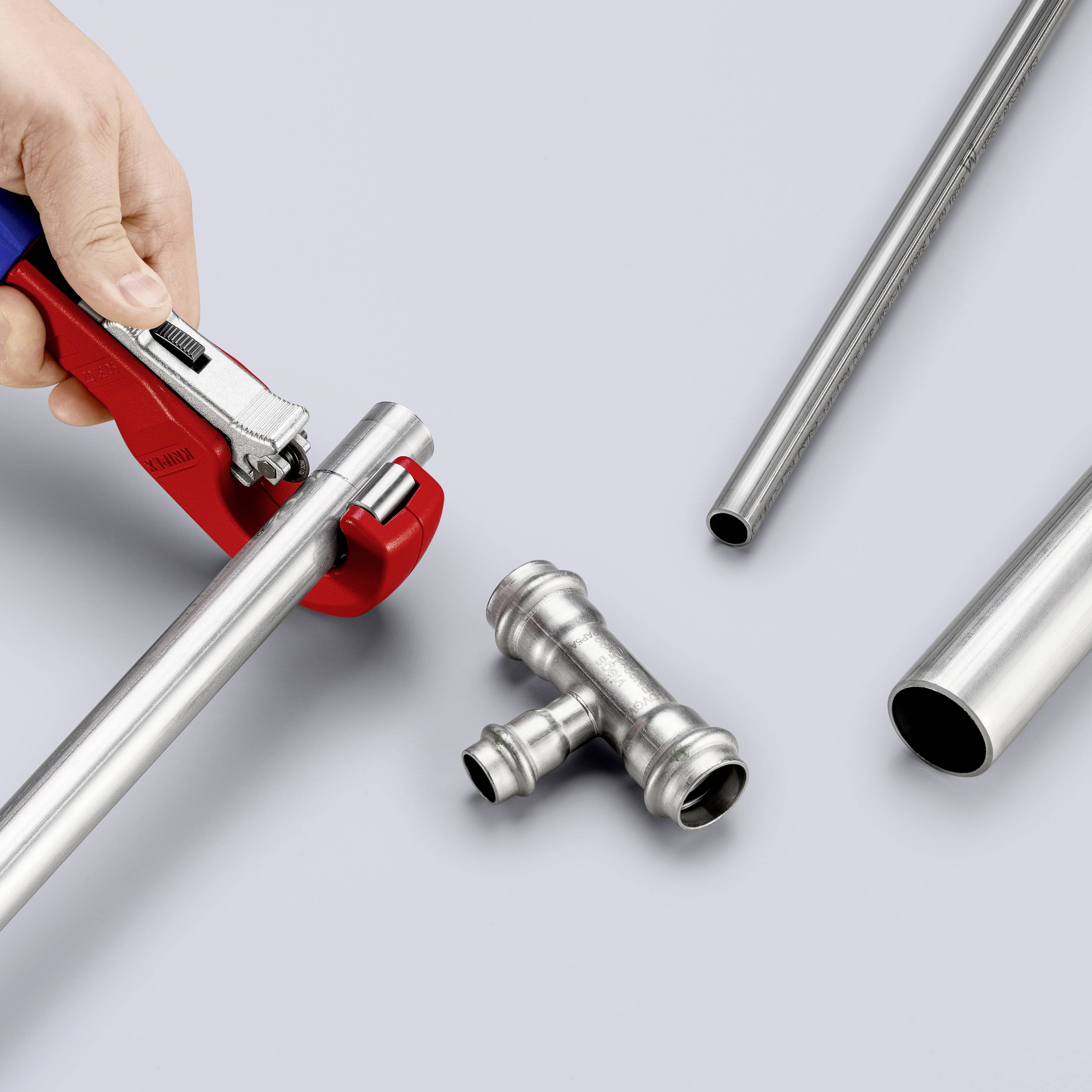 A person is cutting a metal pipe with a pipe cutter. Additional pipe sections and a T-joint are lying on the table.