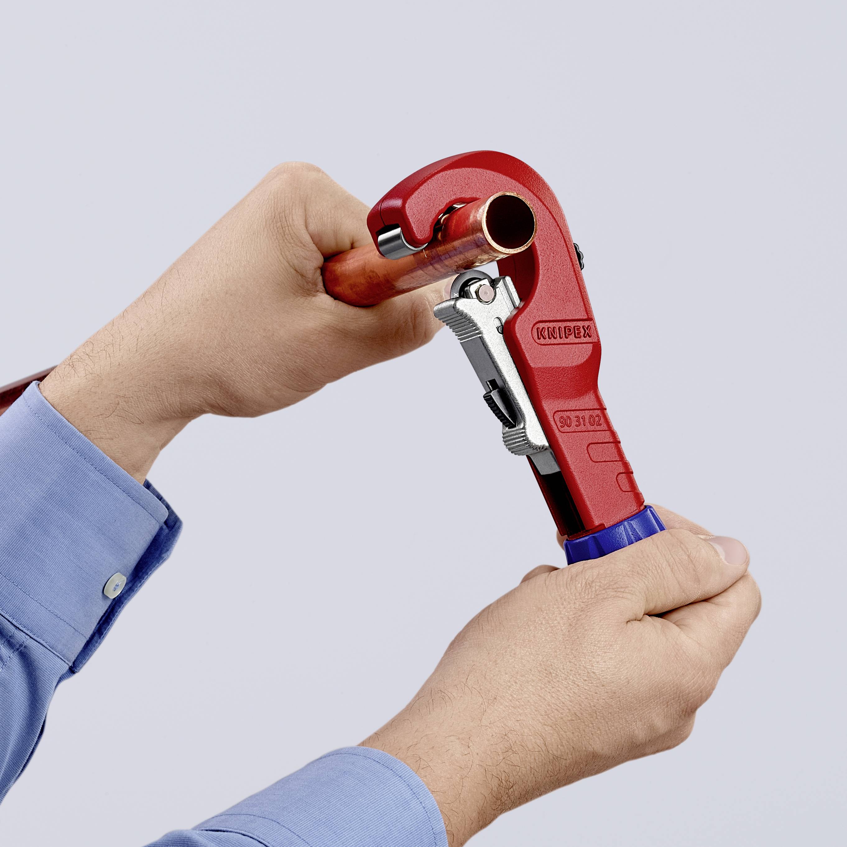 Two hands are holding a red pipe cutter that is cutting a copper pipe. The scene depicts a manual craft activity.