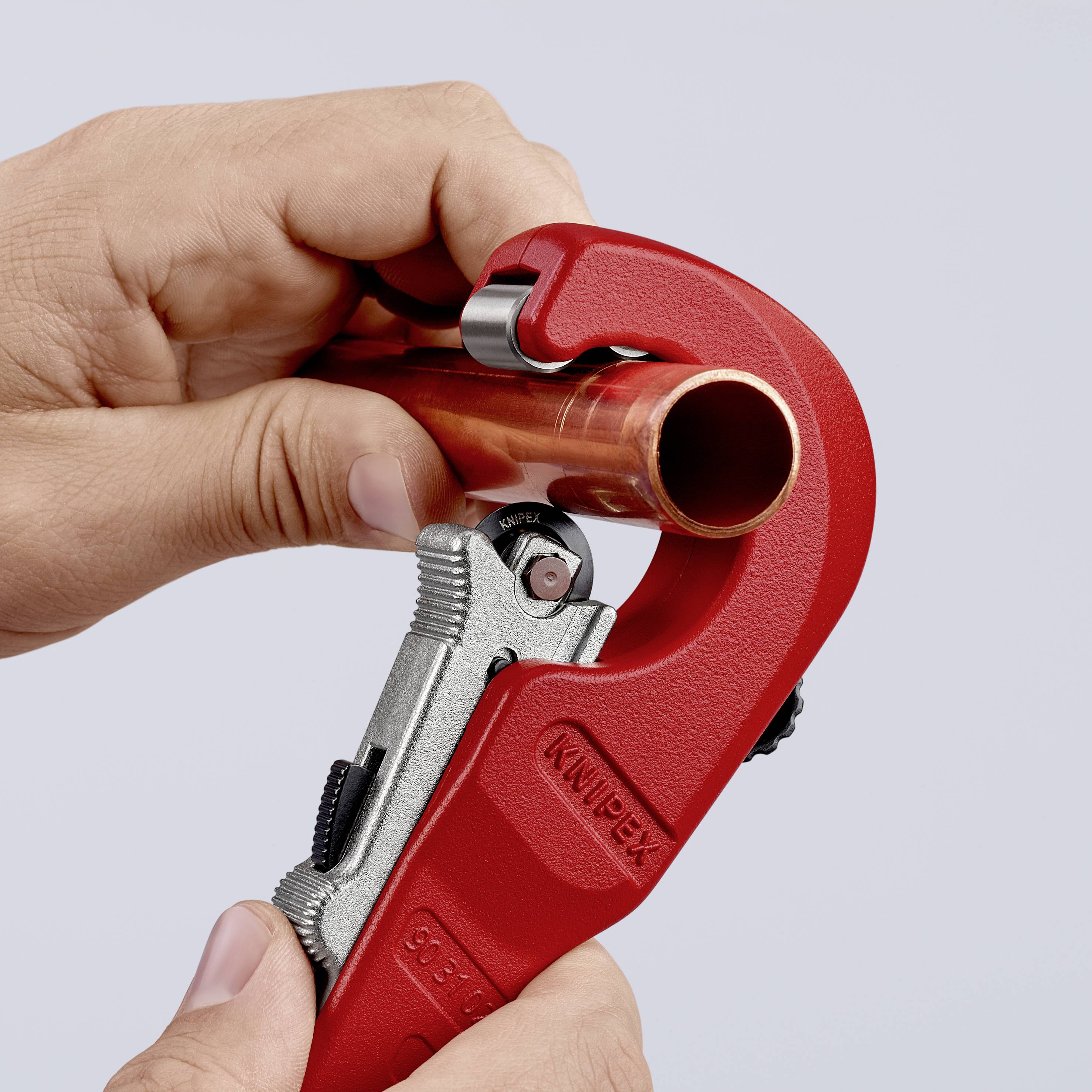 A person is cutting a copper pipe with a red pipe cutter. The pipe cutter has a handle and a sharp cutting wheel.