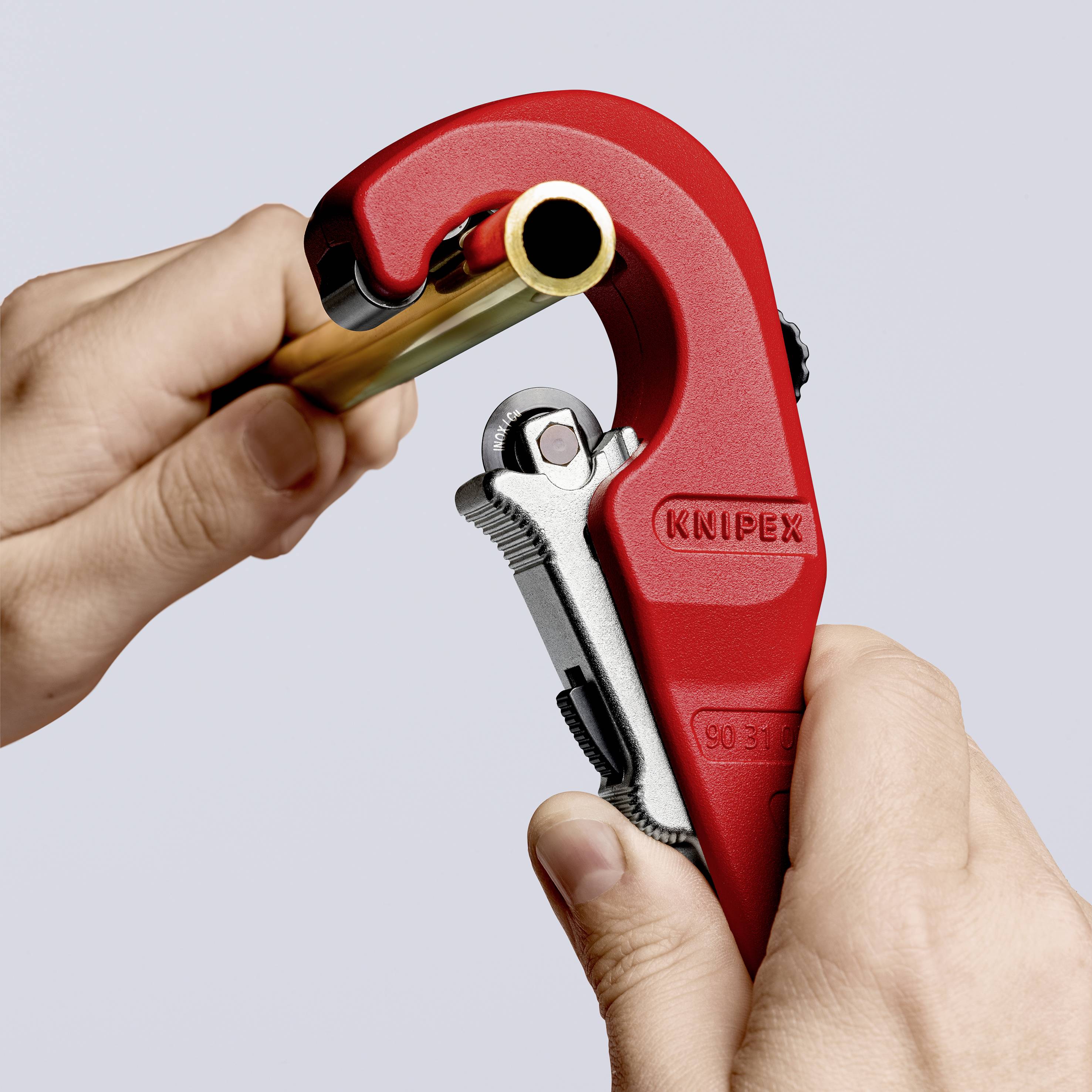 Two hands are holding and using a pipe cutter to cut a copper pipe. The pipe cutter is red and metallic.
