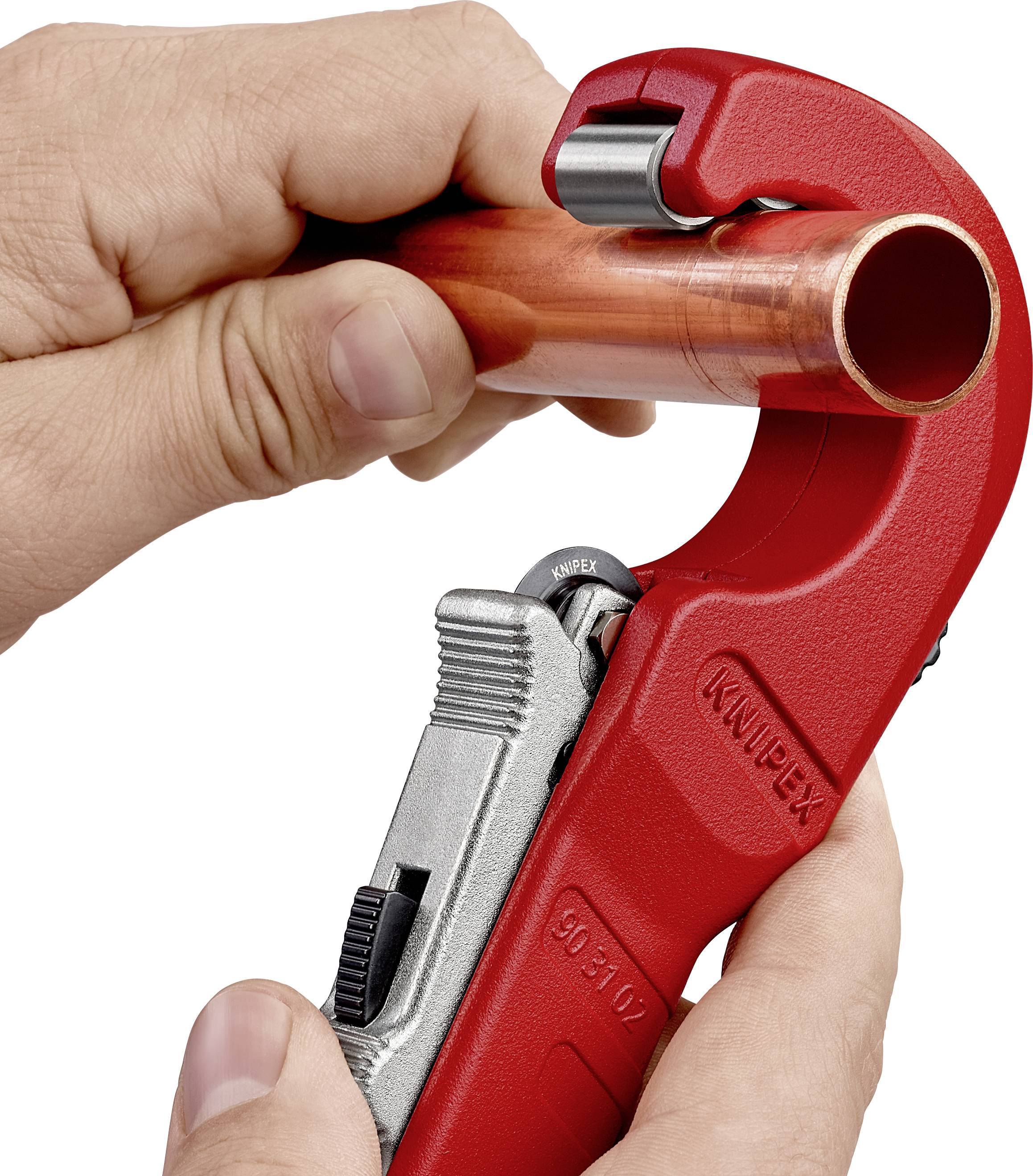 A hand is holding a red pipe cutting tool, cutting through a copper pipe.