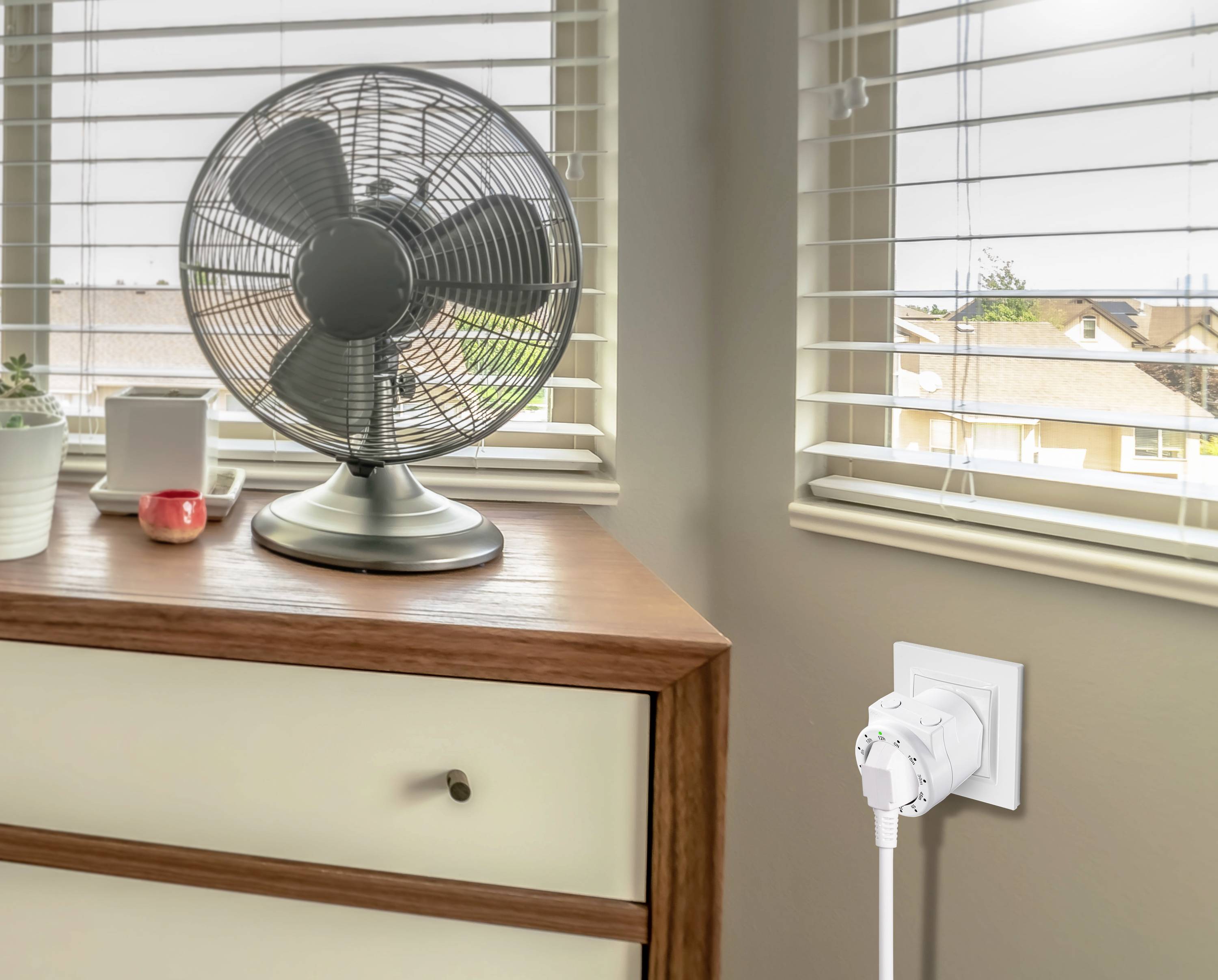 A desk fan sits on a chest of drawers. In the background, windows with blinds can be seen, allowing daylight to enter.