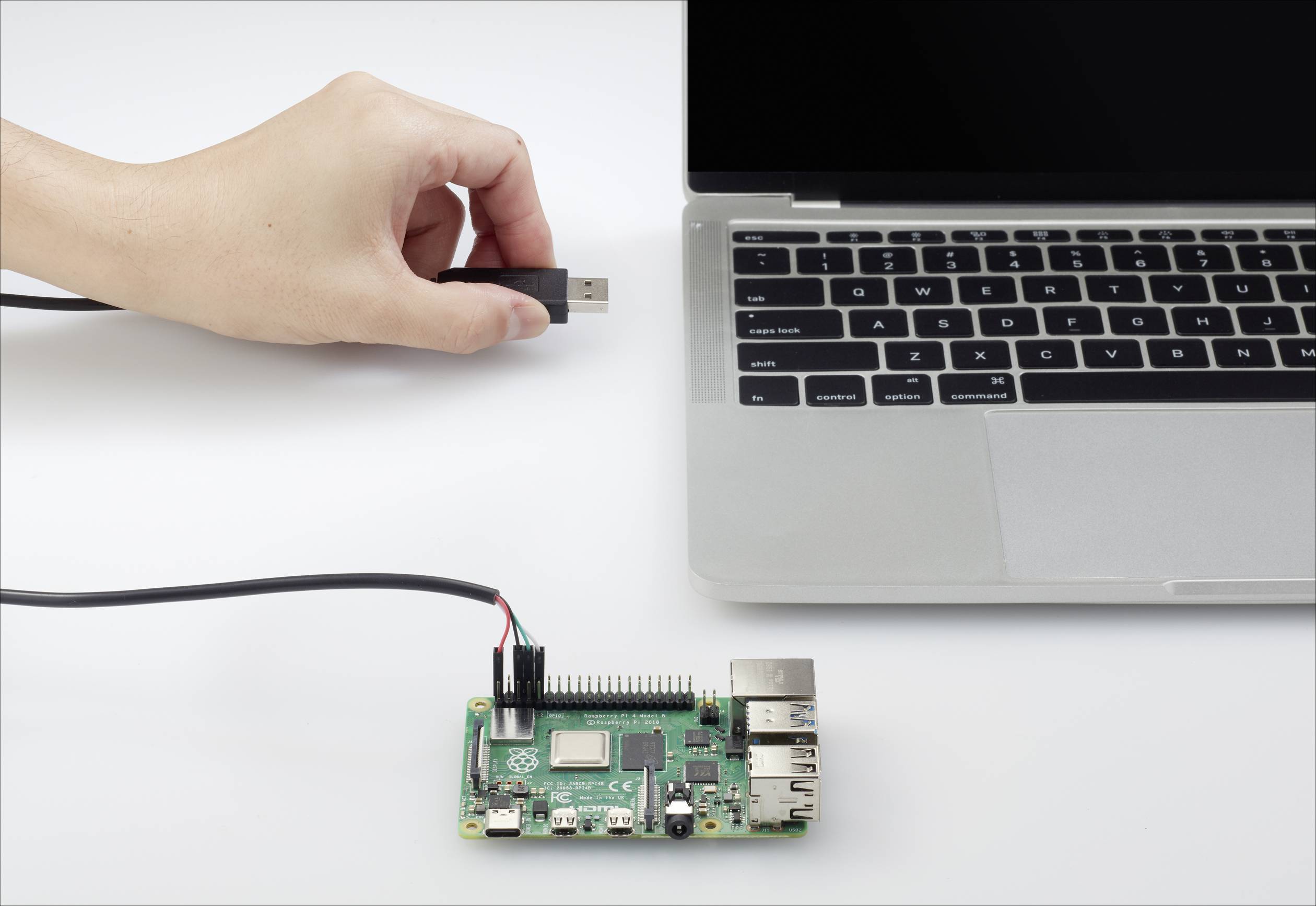 A hand is holding a USB plug next to a laptop. A Raspberry Pi is connected via a cable and is lying on a table.