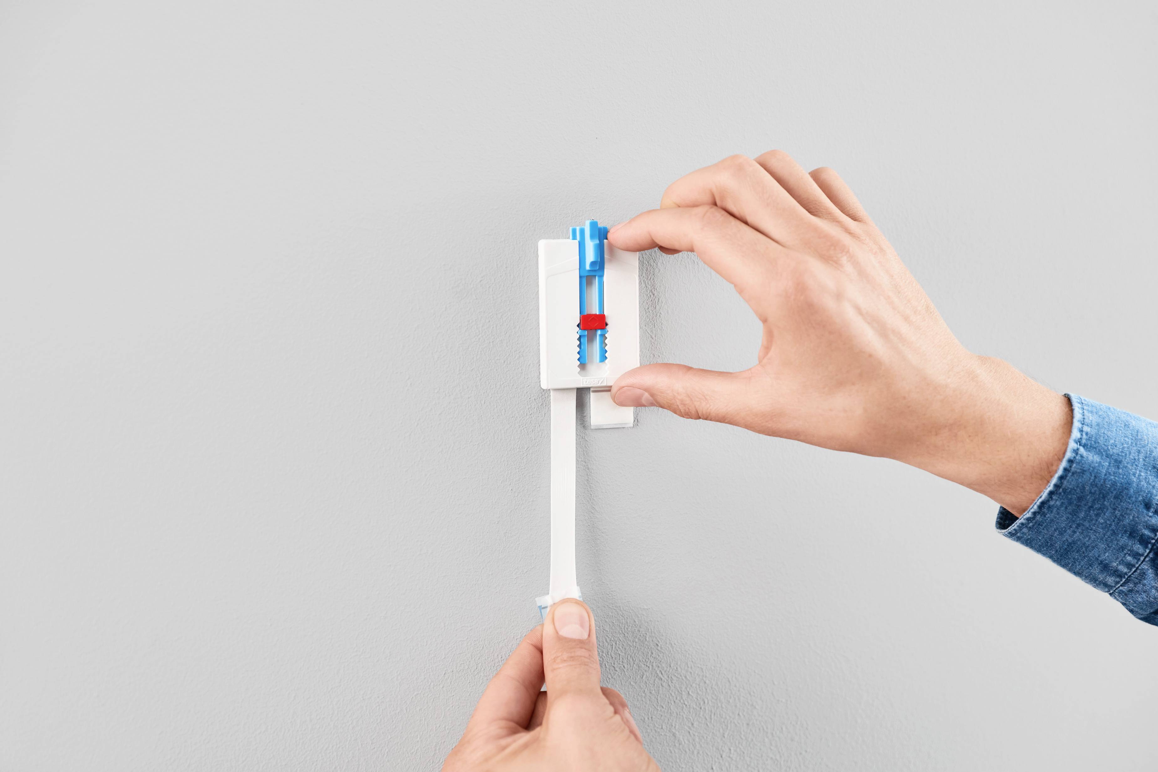 A person is securing a blue and red wall anchor device to a grey wall with their hands.