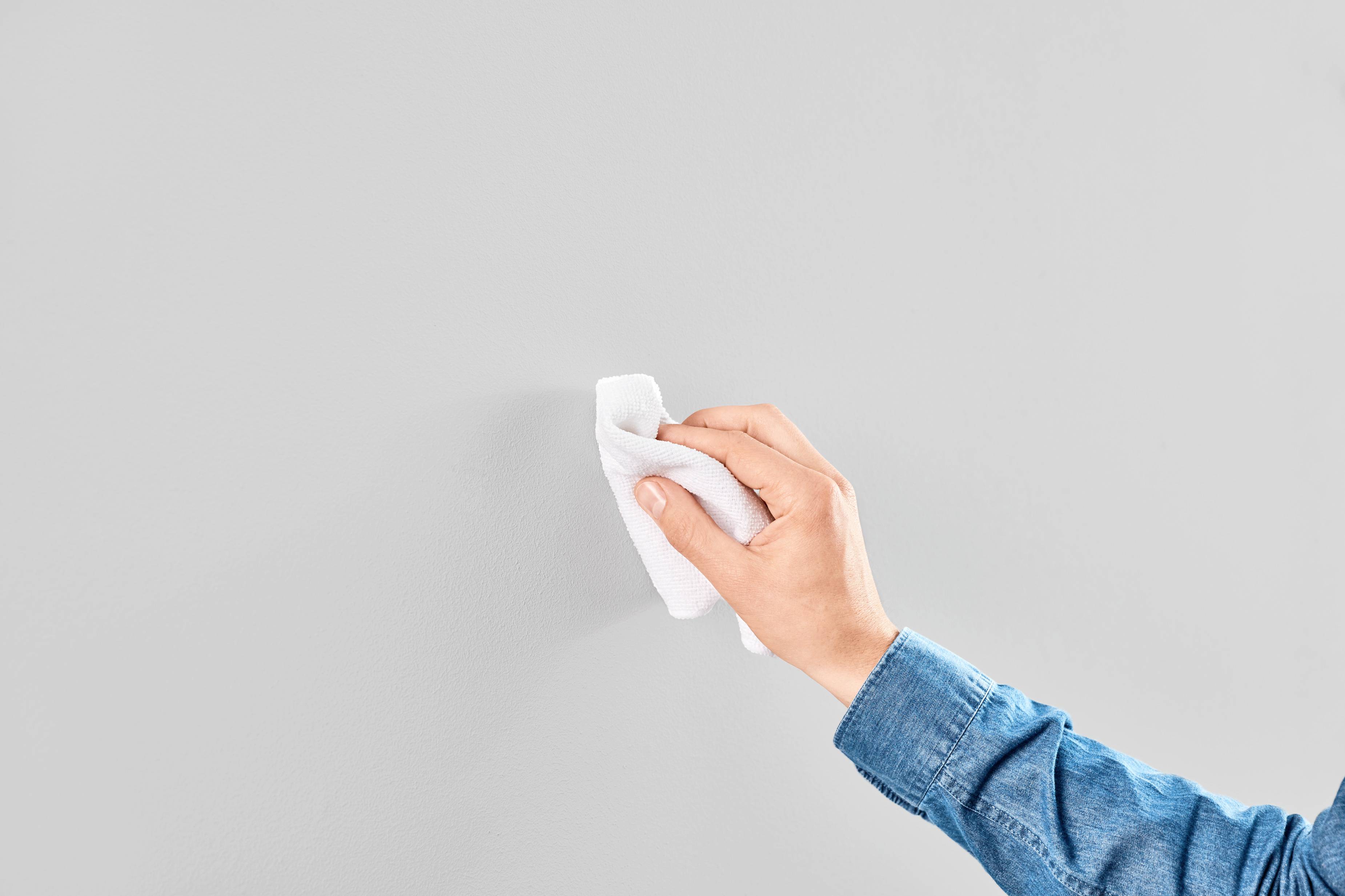 A hand wipes a grey wall clean with a white cloth. The person is wearing a blue shirt.