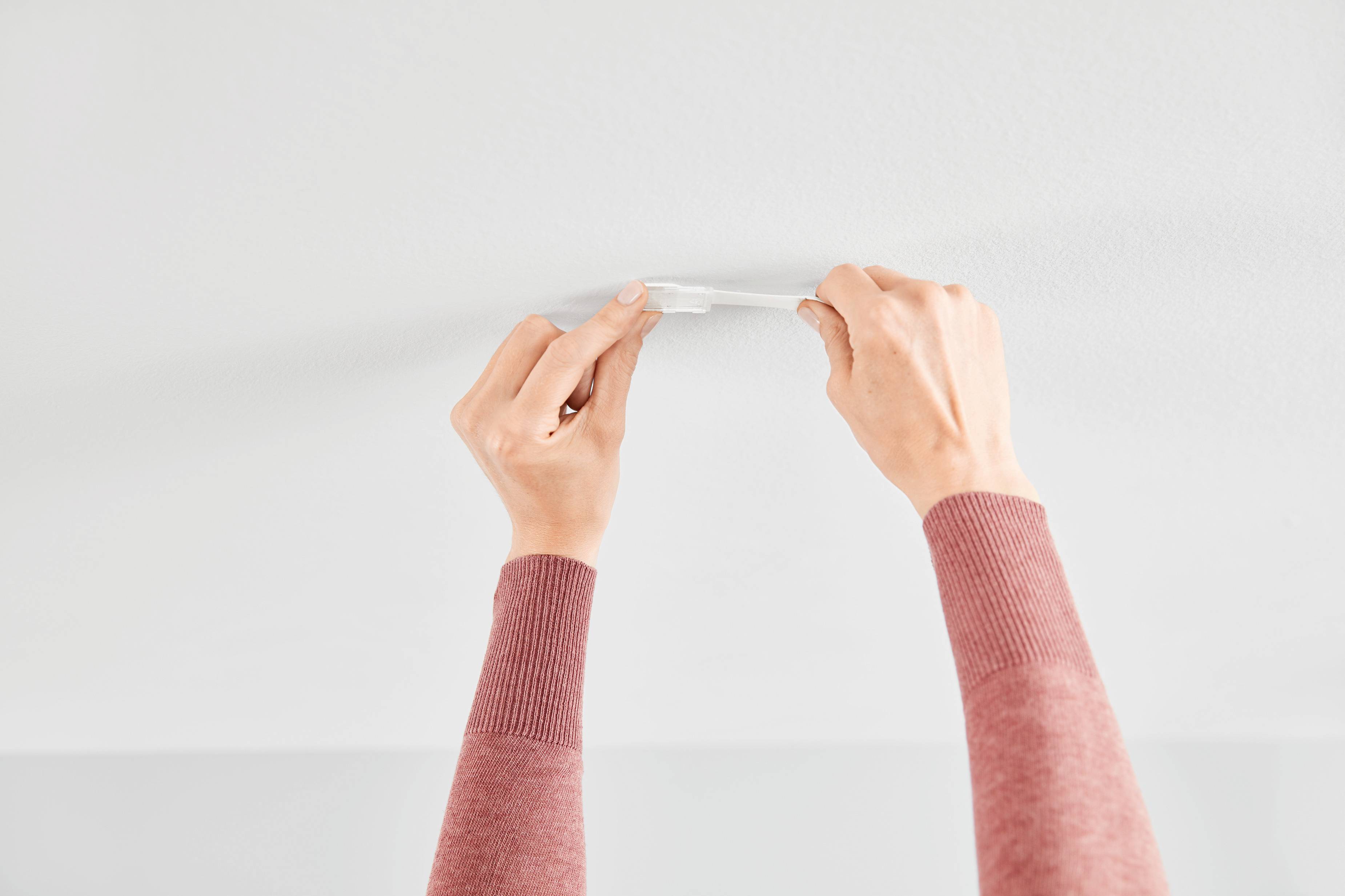 Two hands are sticking transparent tape to the white ceiling. The person is wearing a long-sleeved, pink jumper.
