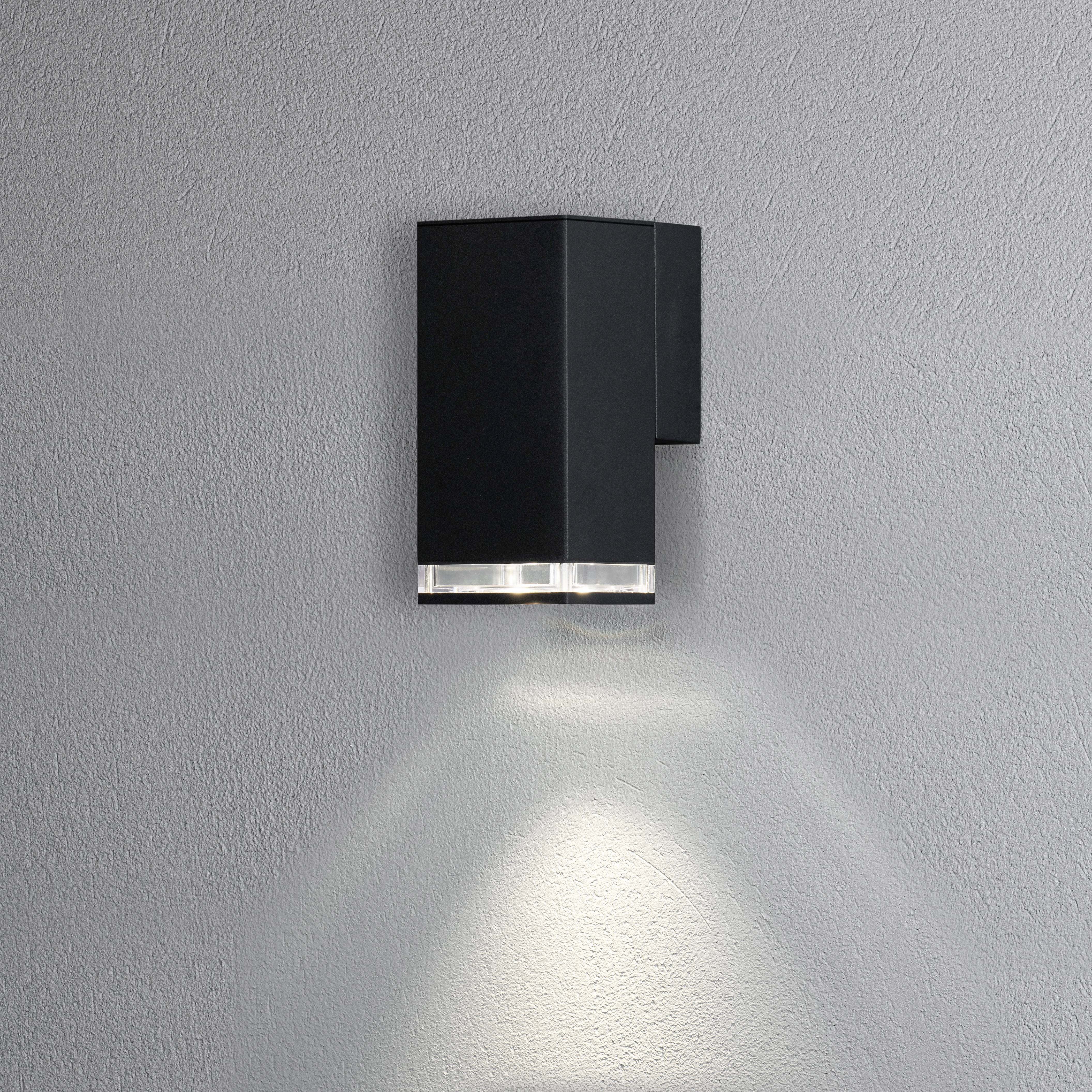 Wall light in a modern, cubic design with a black housing, illuminating downwards and creating soft light on a grey wall.