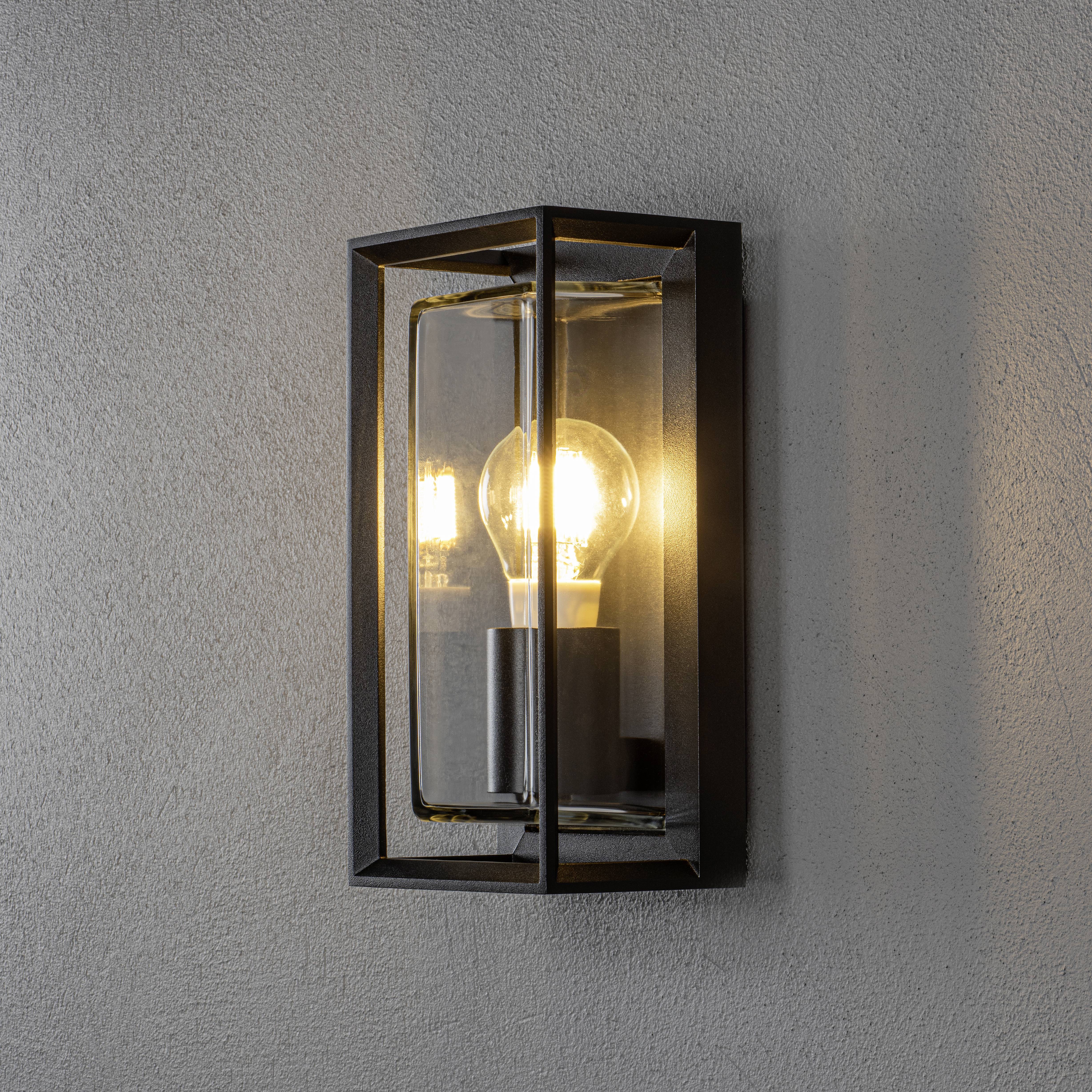 Wall lamp with clear light bulb in rectangular glass box against grey wall, emitting warm light.
