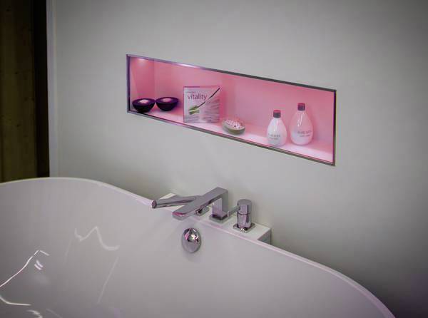 Modern bathroom area with a freestanding white bathtub. An illuminated alcove on the wall displays toiletry products.