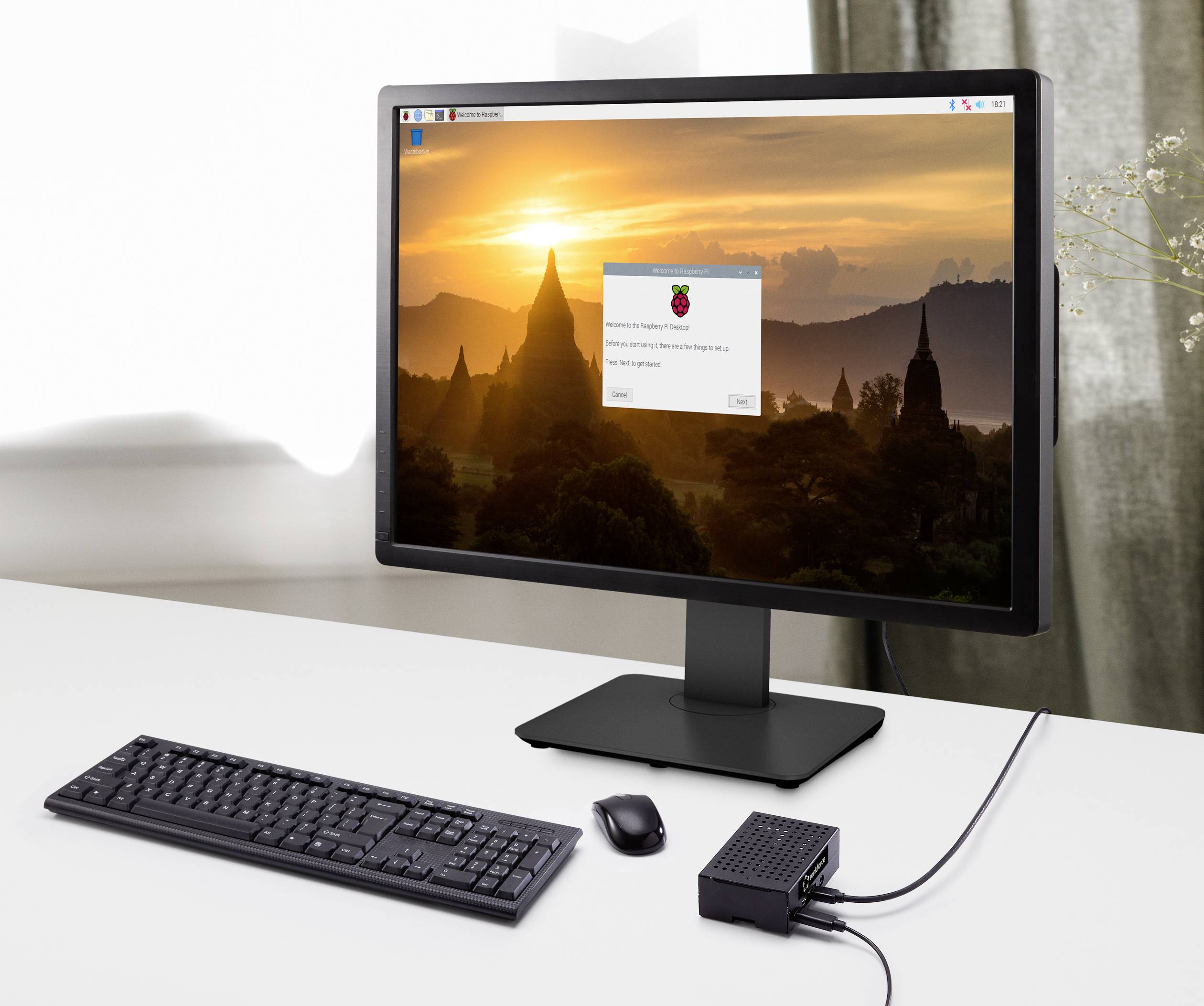Computer screen displaying a landscape and Raspberry Pi operating system logo, keyboard, mouse, and Raspberry Pi device on a white table.