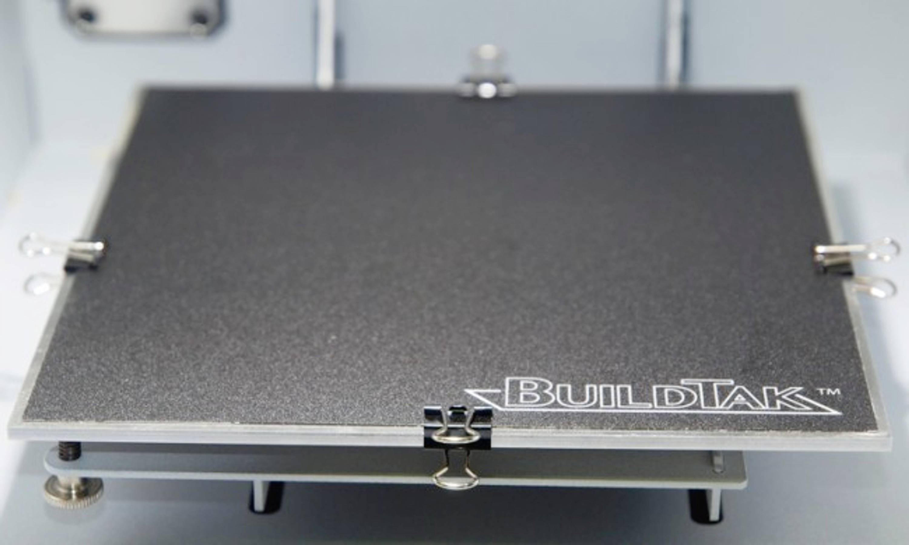 A 3D printer print bed with the inscription 'BuildTak', secured with metal clips.