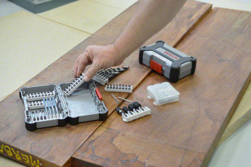 A hand reaches for a drill bit set on a wooden table. The boxes are open, and various drill bit attachments are visible.