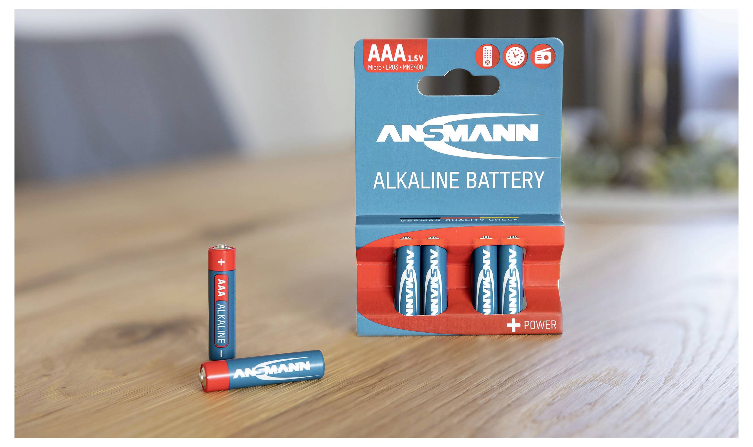 A pack of three blue Ansmann alkaline batteries, with one battery standing separately beside the pack on a wooden table.